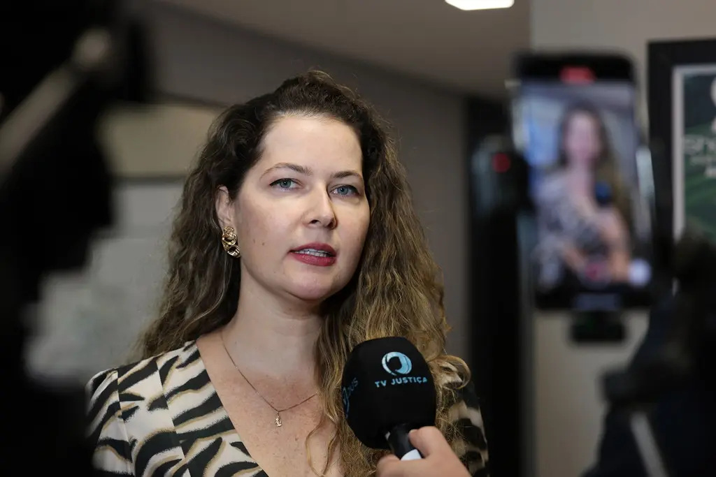 Foto horizontal colorida, em plano fechado, que mostra a secretária-chefe do Gabinete de Enfrentamento à Violência contra a Mulher, delegada Mariell Antonini durante entrevista à TV Justiça. Ela é uma mulher branca, de olhos azuis, com cabelos longos, loiros e cacheados, usando blusa estampada.