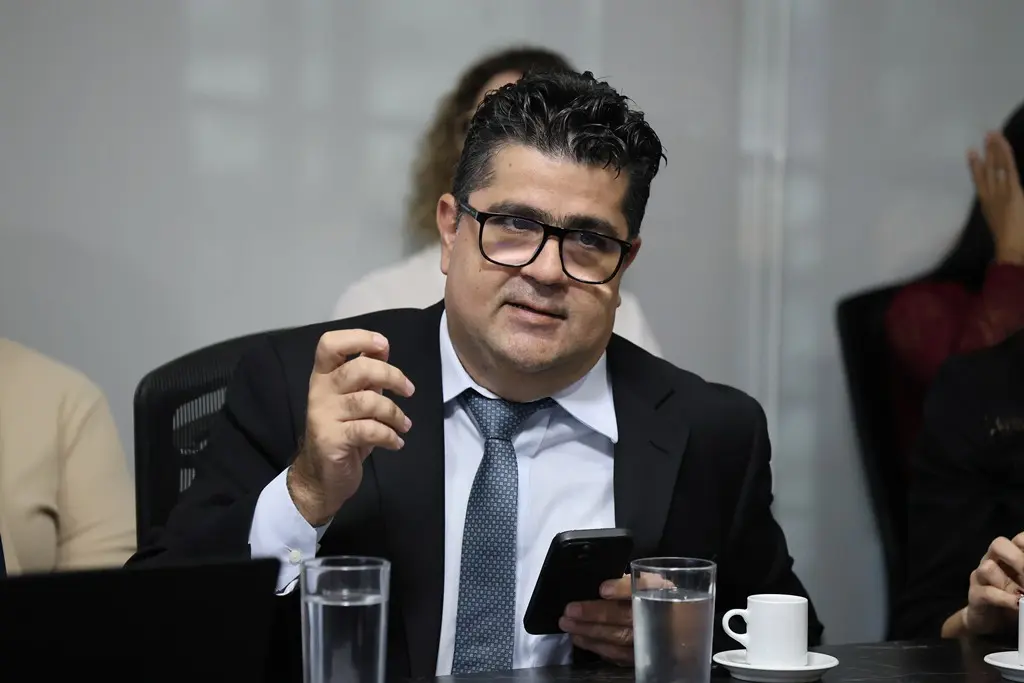 Foto horizontal colorida, em plano fechado, que mostra o juiz Marcos Terêncio, sentado, falando durante uma reunião. Ele é um homem branco, de olhos e cabelos escuros, usando camisa branca, gravata cinza e terno preto.