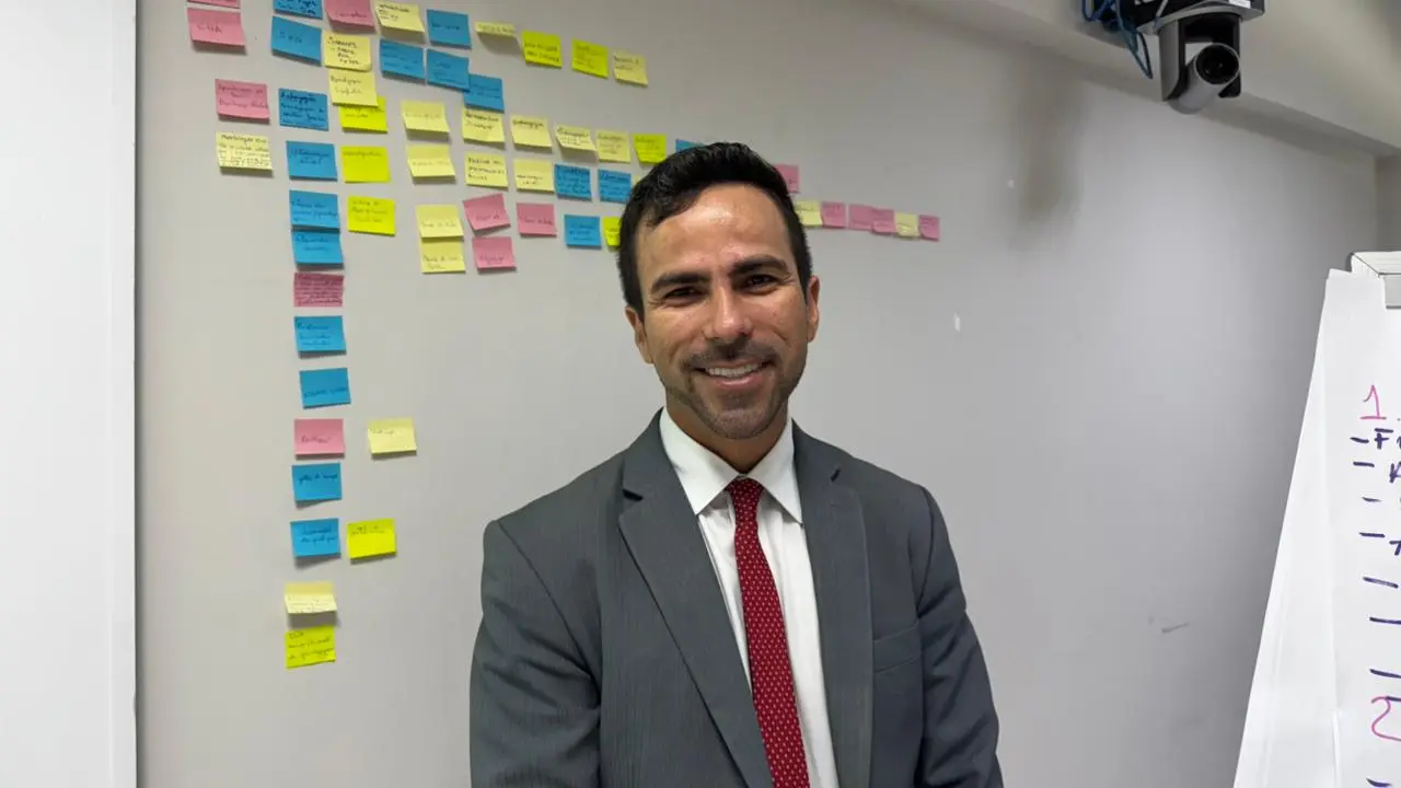 Homem branco sorridente de cabelos curtos escuros, vestindo terno cinza, camisa branca e gravata vermelha, posa à frente de uma parede branca. Ao fundo, diversos post-its coloridos estão organizados em colunas e linhas, indicando um painel de planejamento ou lousa de atividades.