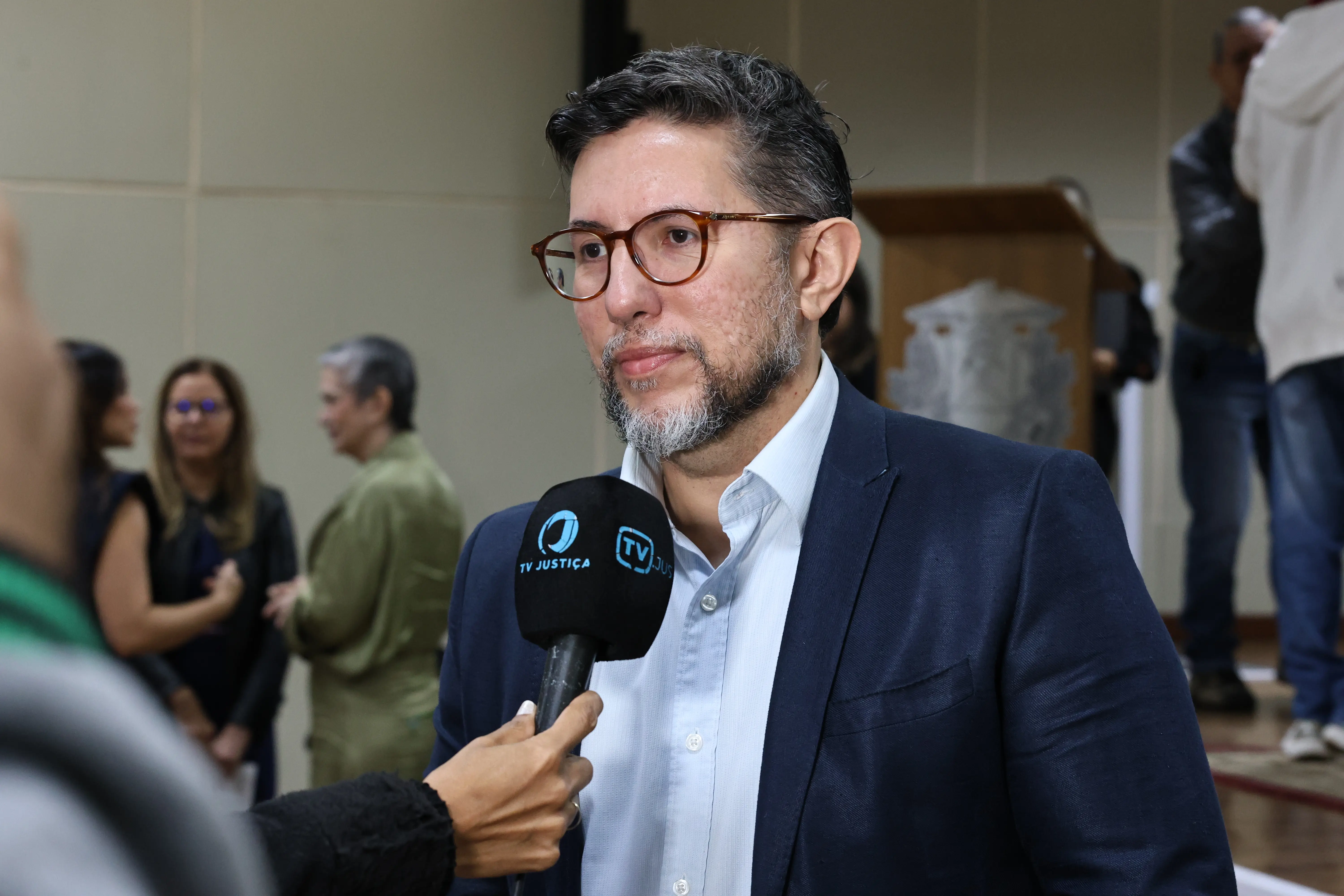 Homem branco, com cabelo preto e barba grisalha. Usa terno azul marinho, com camisa azul claro. Ele fala a um microfone, que tem escrito em azul 