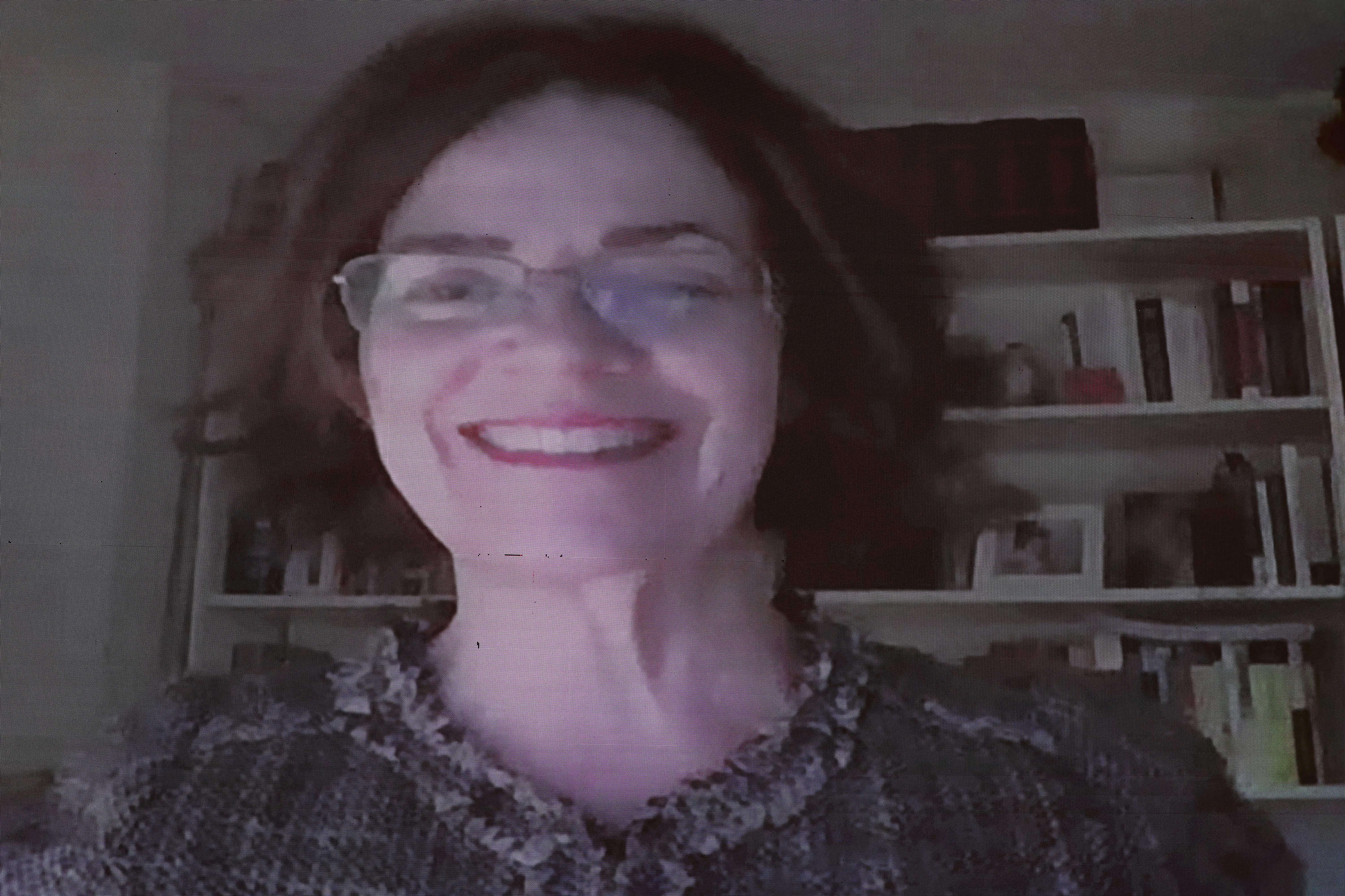 Close-up digital e pixelado do rosto de uma mulher sorrindo. Ela tem cabelos escuros, usa óculos de grau e aparece em uma transmissão de vídeo.