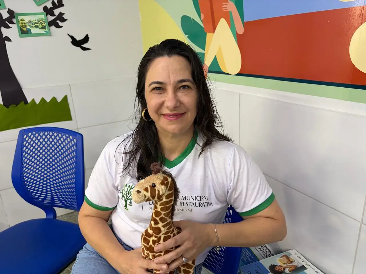 Mulher sentada sorri segurando uma girafa de pelúcia. Ela usa camiseta branca com detalhes verdes onde se lê