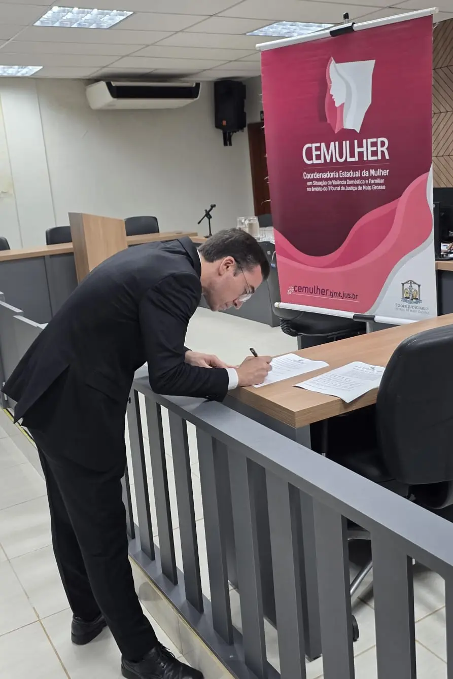 Homem de terno preto assina documentos sobre balcão de madeira. Ao fundo, banner rosa da CEMULHER — Coordenadoria Estadual da Mulher do Tribunal de Justiça de Mato Grosso.