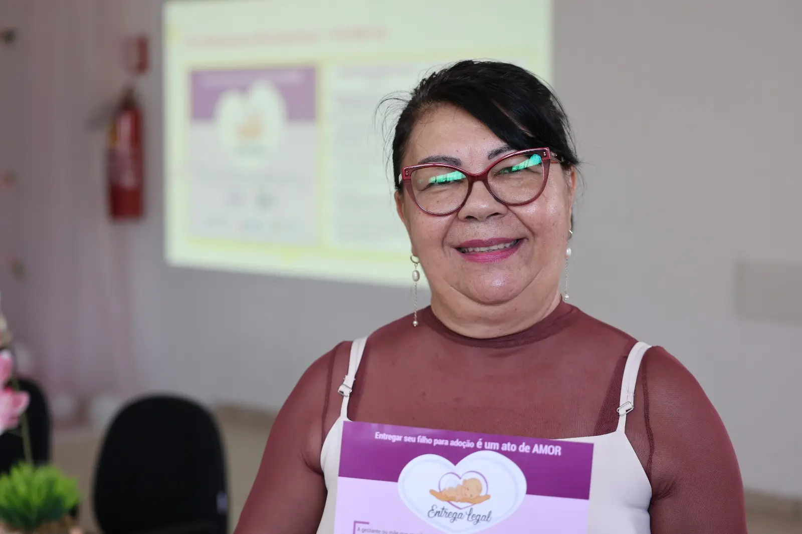 A imagem mostra a assistente social do Cras Dom Aquino, Joanil Maria Casarotto, segurando um folder da campanha Entrega Legal. Ela usa blusa em tom vinho transparente com alças claras e óculos de armação vermelha. 