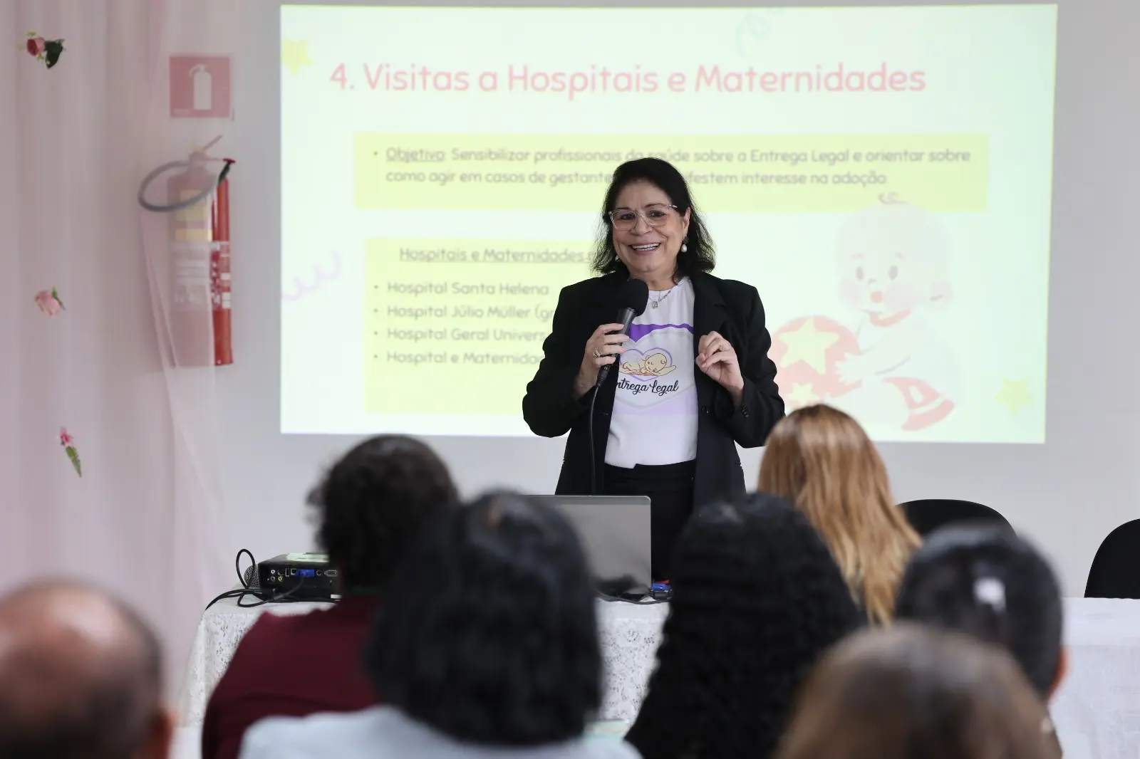 A imagem mostra a juíza Gleide Bispo Santos durante palestra da Semana Estadual de Conscientização sobre a Entrega Voluntária. Ela segura um microfone e sorri enquanto fala ao público. 