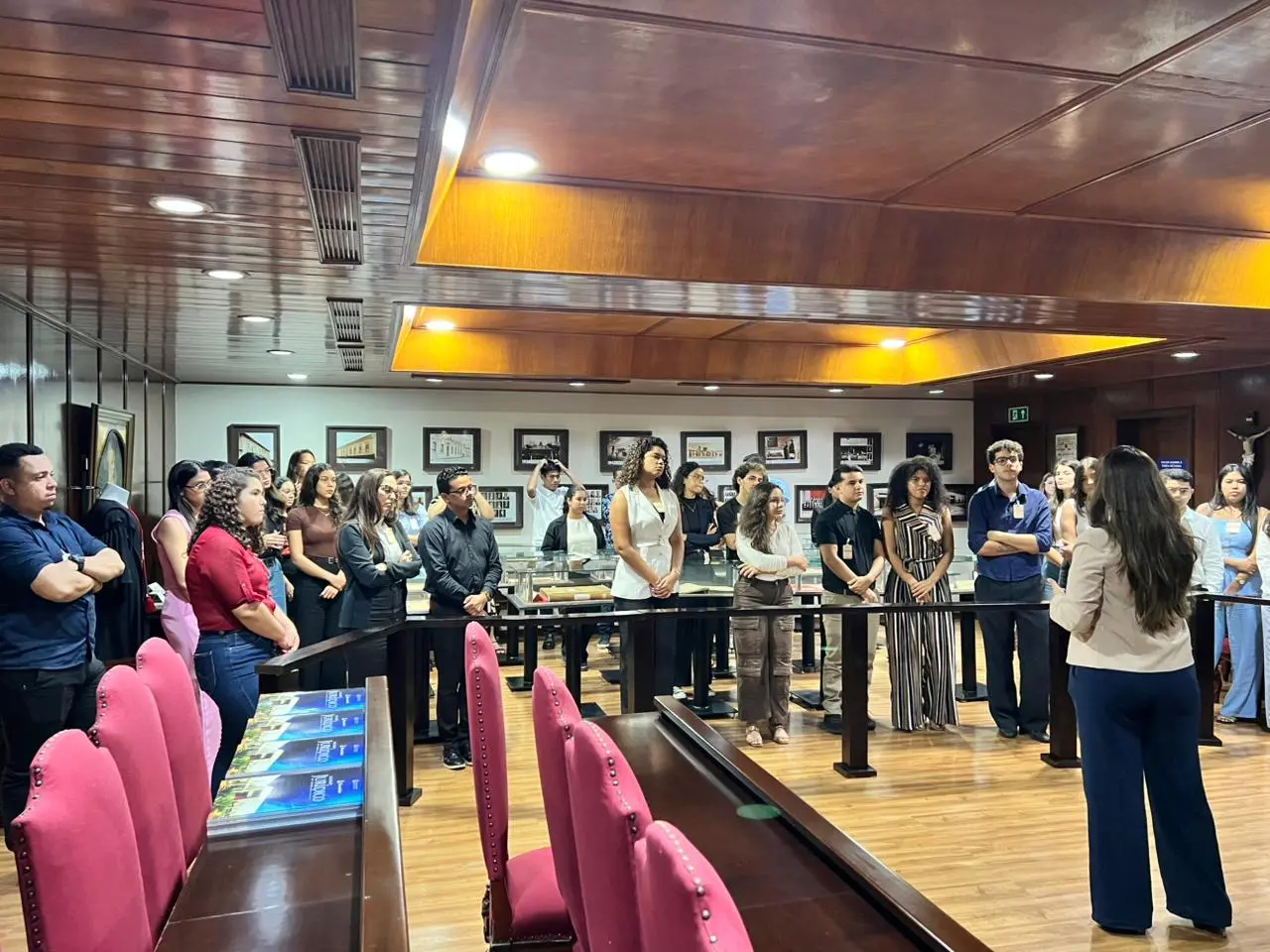 Grupo de jovens está reunido em semicírculo em sala institucional, ouvindo uma mulher de costas que conduz uma apresentação. Sobre a mesa, exemplares do Glossário Jurídico.