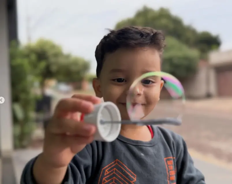 Menino sorrindo em primeiro plano segura um bastão de bolha de sabão. Há uma bolha transparente inflada na ponta do arco. O fundo mostra árvores e uma rua residencial desfocadas.