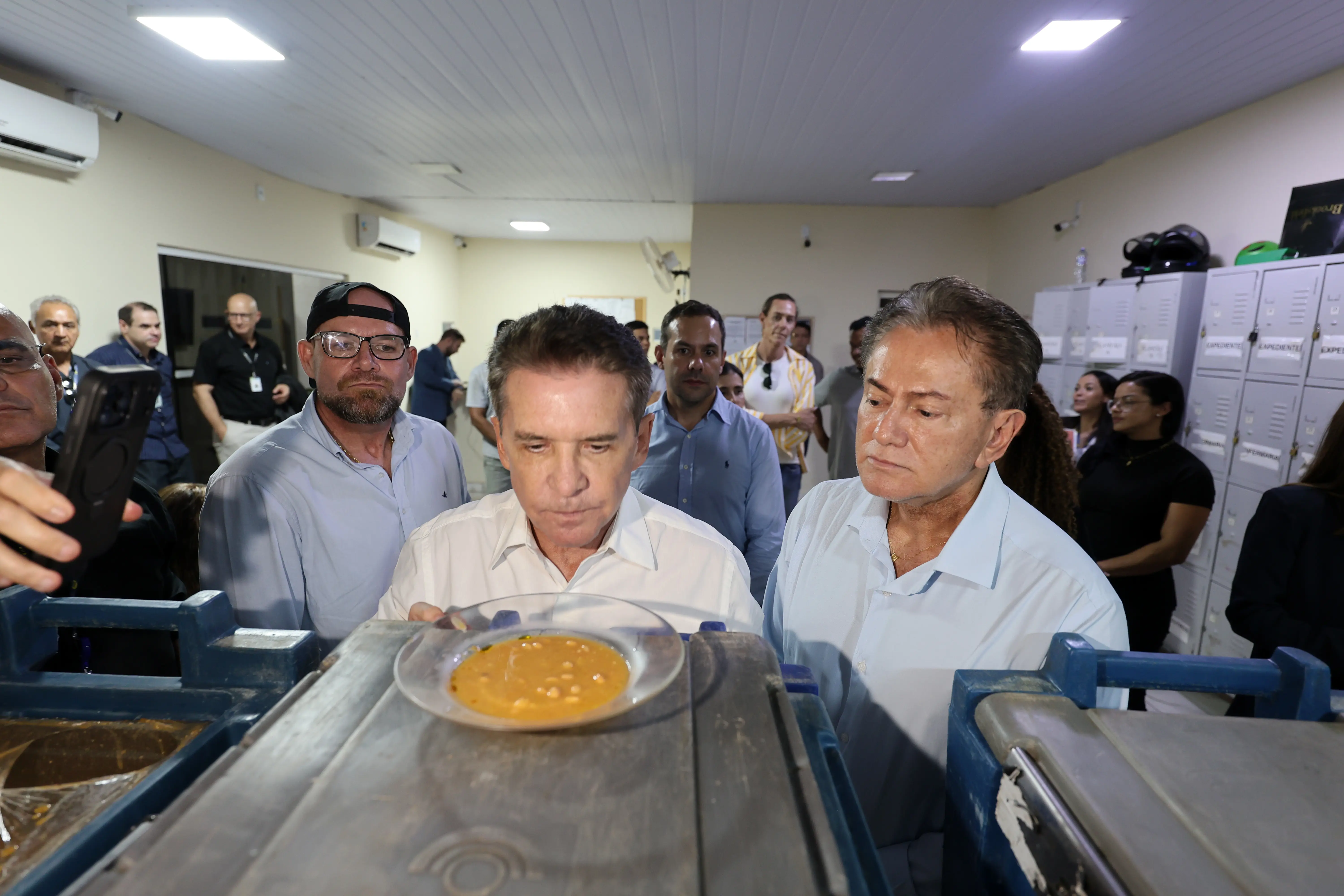Dois homens de camisa branca se debruçam sobre um prato com comida amarelada sobre bandeja metálica. Ao fundo, grupo de pessoas observa a cena em ambiente fechado.