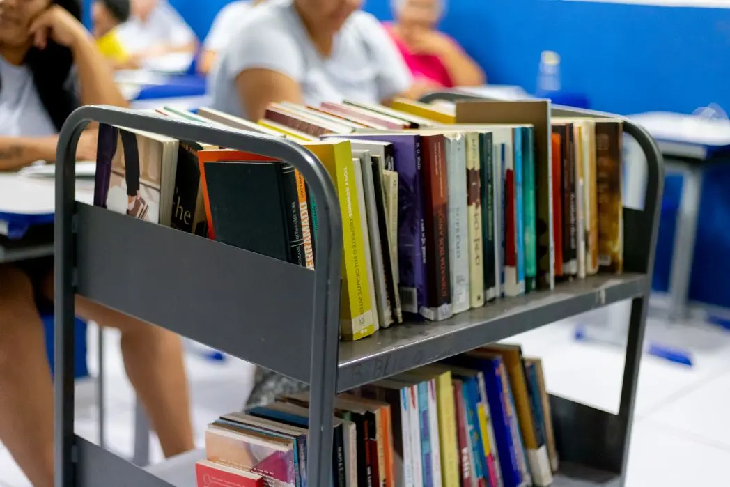 Foto horizontal que mostra um carrinho cheio de livros, em primeiro plano. Em segundo plano, desfocado, aparece a sala de aula com mulheres privadas de liberdade sentadas nas carteiras. 