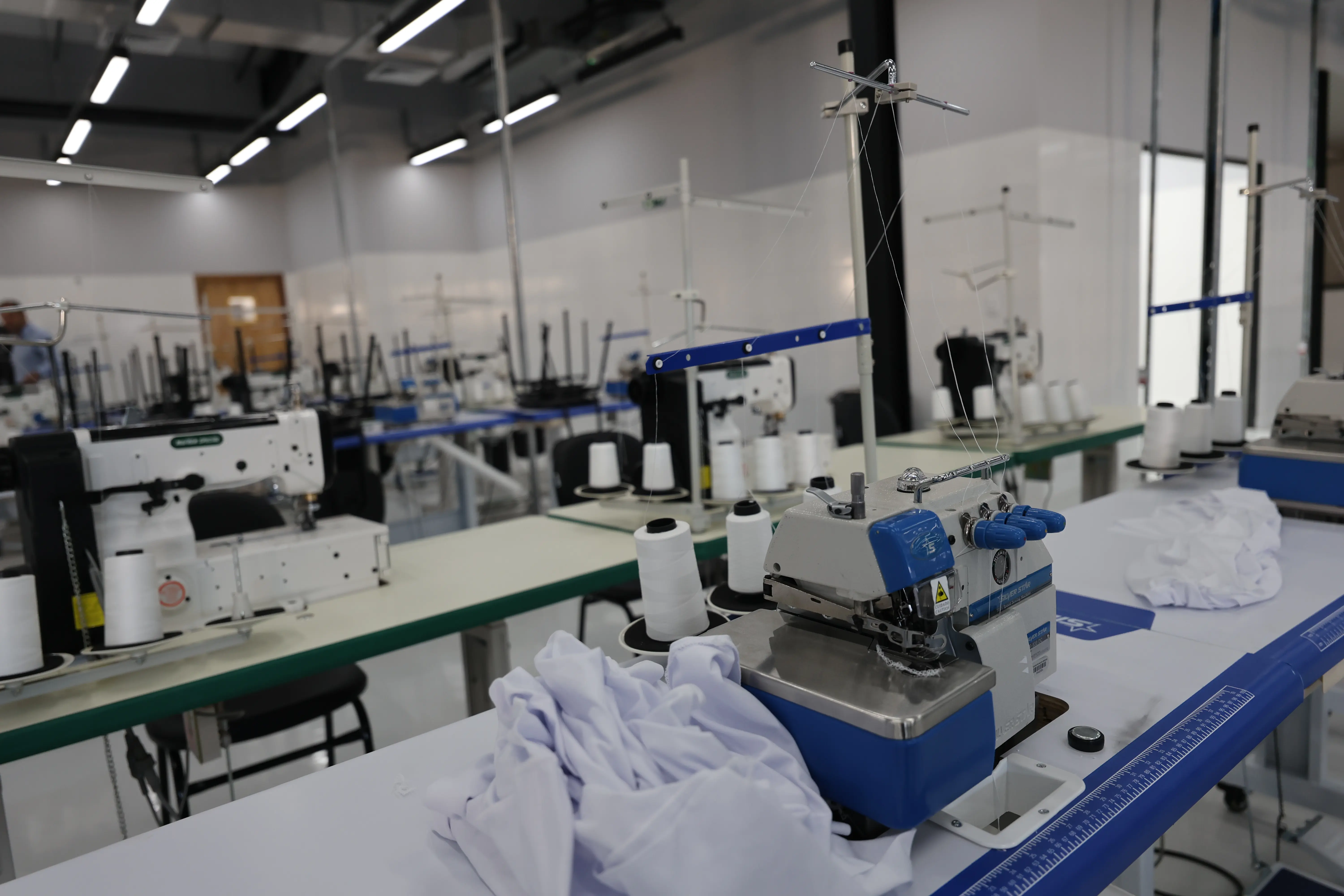 Foto em plano aberto de um ateliê de costura, com máquinas de costura industriais e tecidos brancos compondo a imagem. 