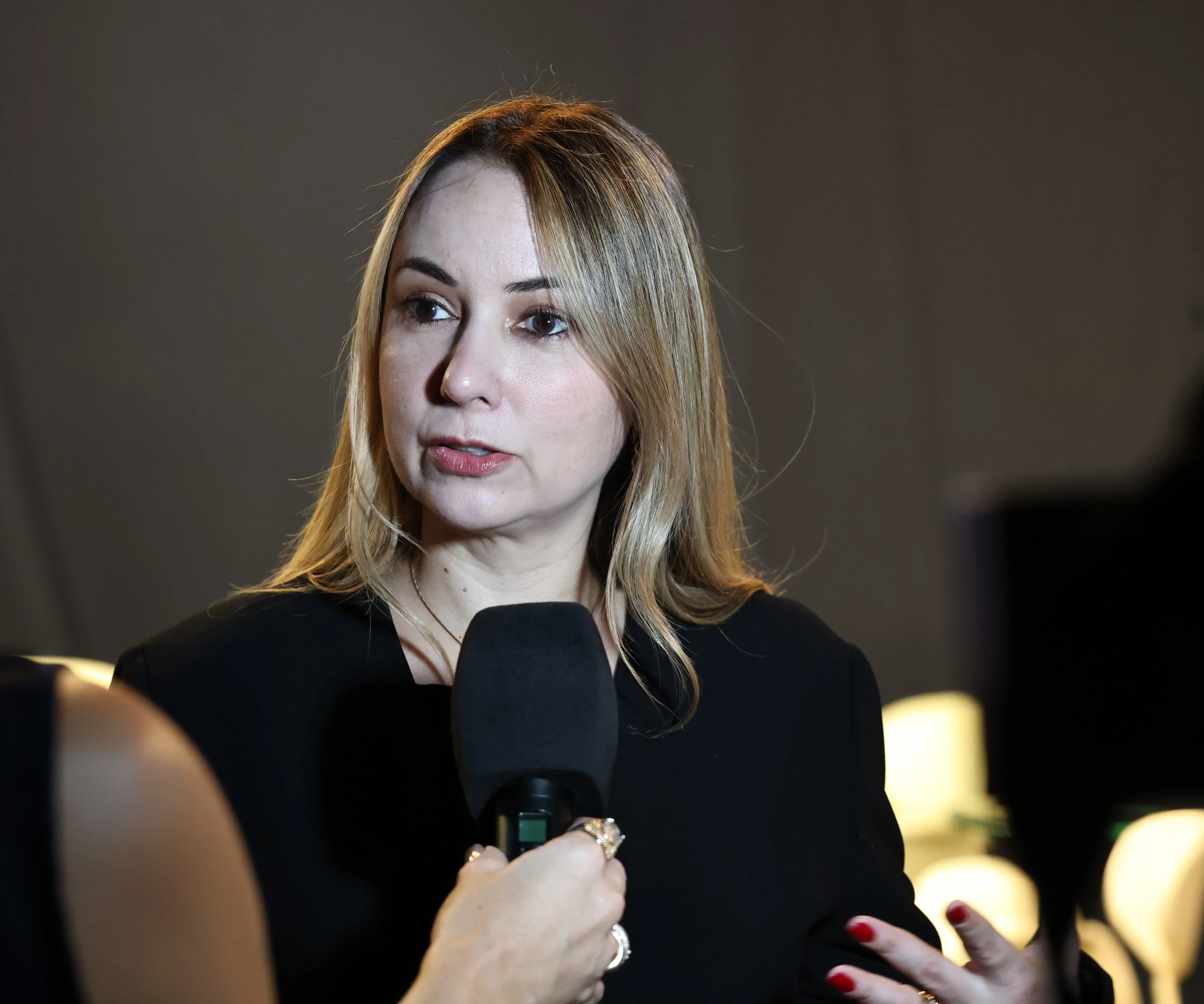 A imagem mostra a juíza Ana Graziela falando ao microfone durante entrevista para a TV Justiça. Ela é uma mulher de pele clara, cabelos lisos e loiros e olhos escuros. Veste roupa preta. 
