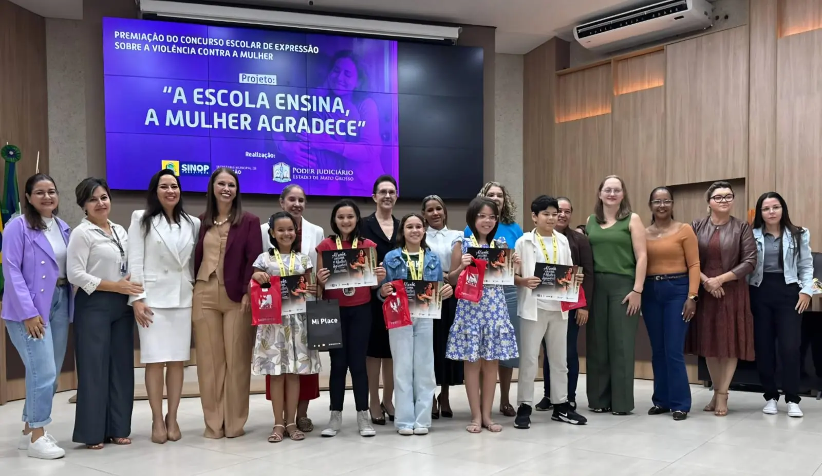 Adultos e crianças posam juntos em um palco. Os estudantes seguram certificados e sacolas de brindes. Ao fundo, um grande telão roxo exibe: