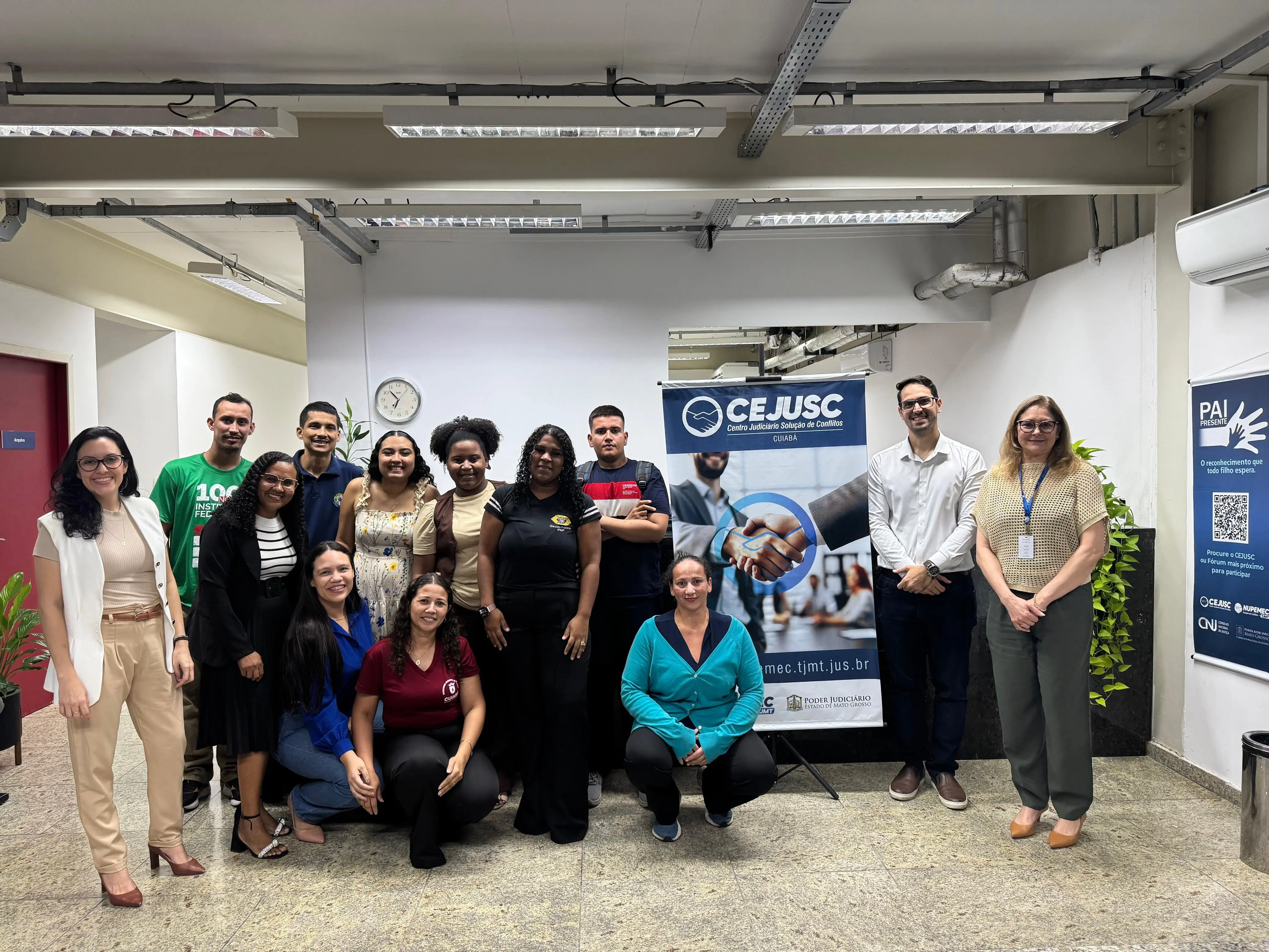 Grupo de pessoas reunidas em um ambiente interno posa sorrindo para foto ao lado de um banner azul do CEJUSC. Alguns estão em pé e duas mulheres estão agachadas à frente.