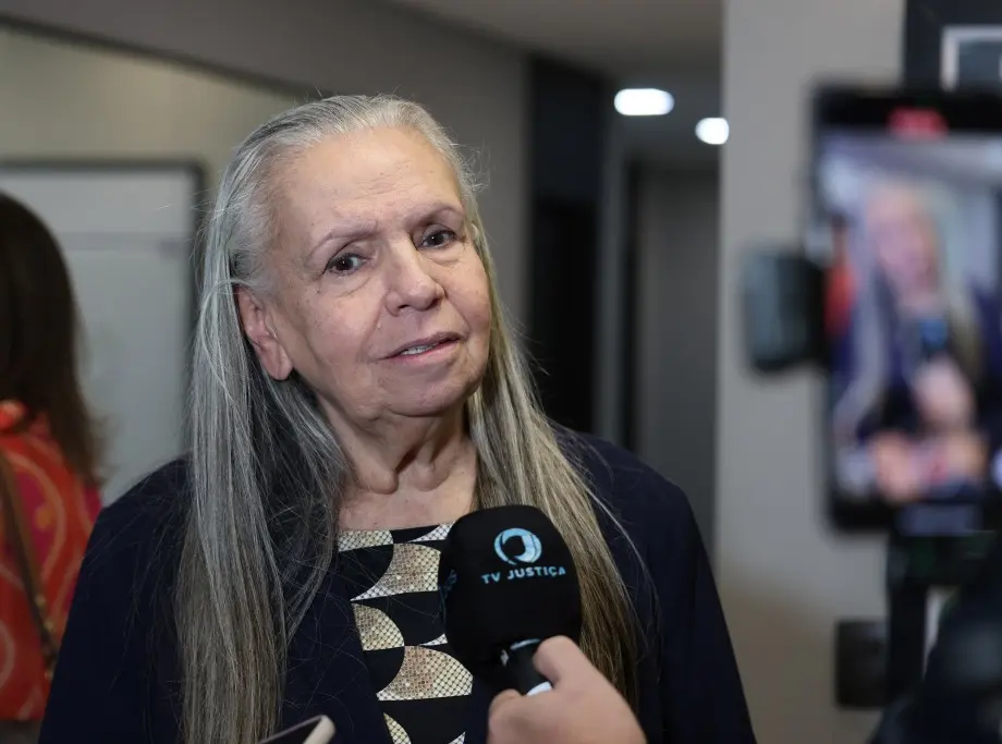 Foto horizontal colorida, em plano fechado, que mostra a desembargadora Maria Erotides Kneip durante entrevista à TV Justiça. Ela é uma senhora branca, de cabelos longos, lisos e grisalhos, usando blusa com estampa geométrica preto e dourado e blazer azul marinho.