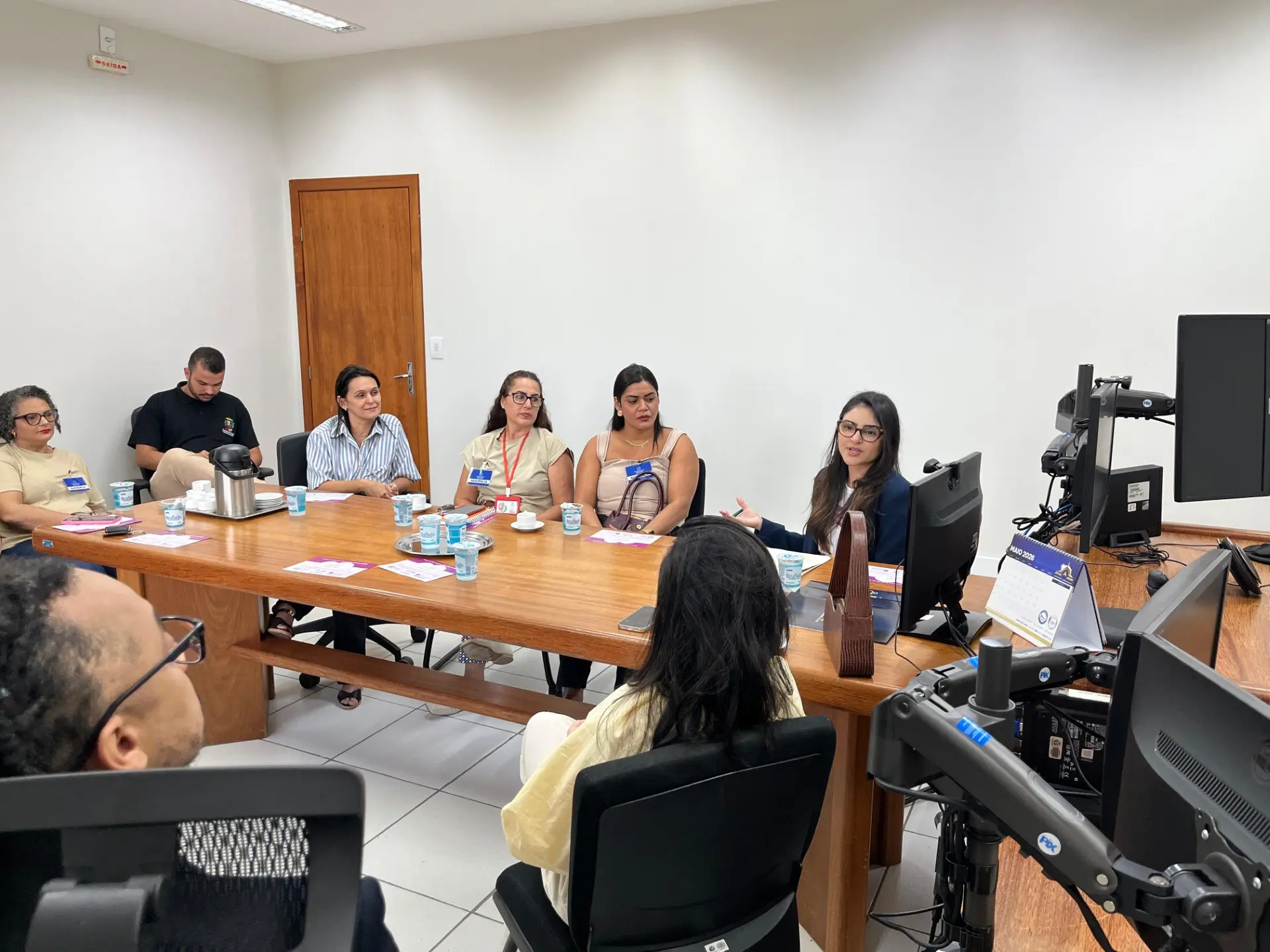 Foto de participantes da roda de conversa sobre entrega voluntária para adoção reunidos em sala de reuniões do Fórum de Juara. Na imagem, uma mulher fala aos demais participantes sentados ao redor de uma mesa de madeira. A sala possui computadores, cadeiras de escritório e iluminação interna.