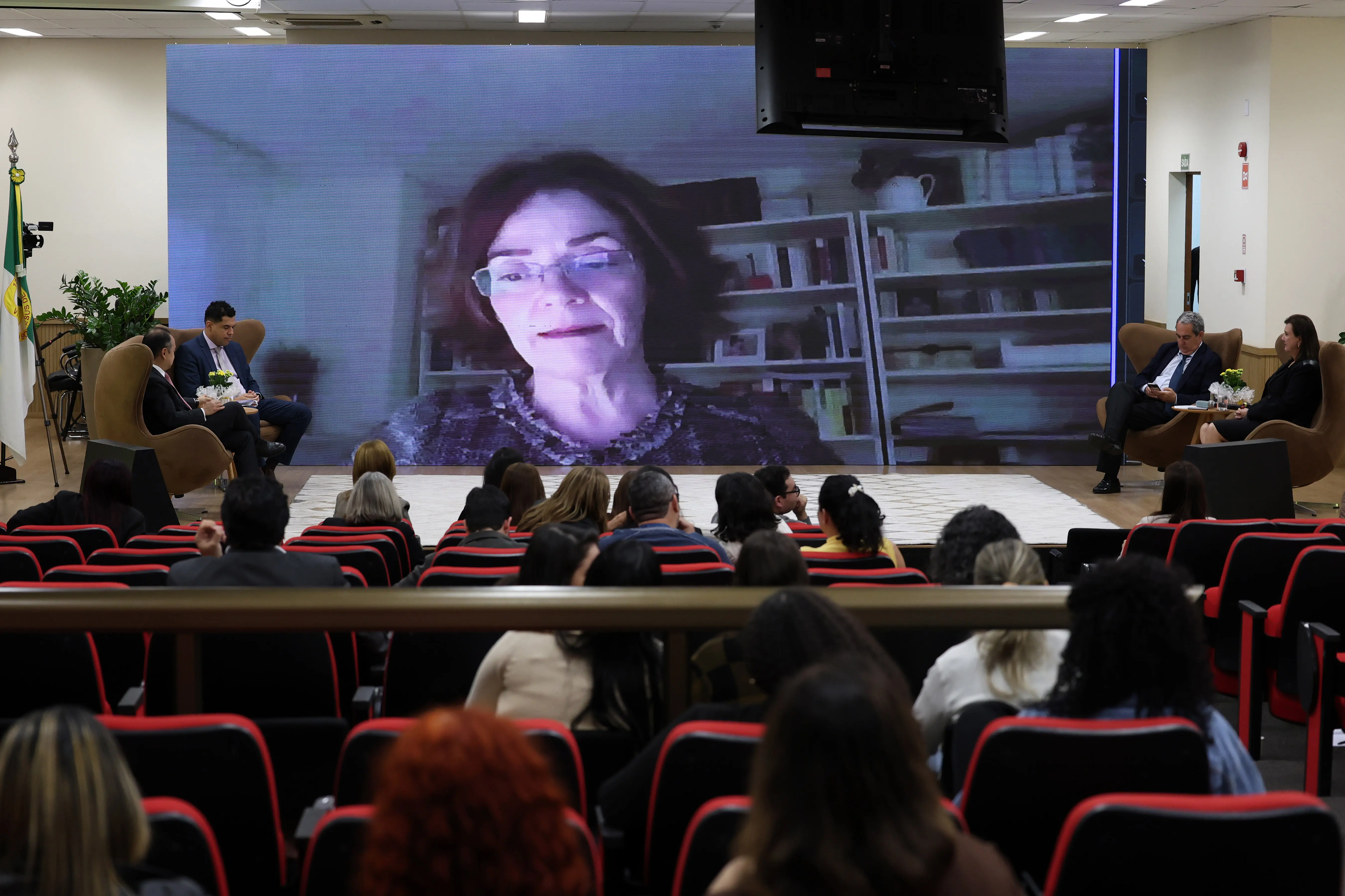 Vista de um auditório com público assistindo a uma palestra. No palco, quatro pessoas em poltronas ladeiam um grande telão que exibe a imagem de uma mulher de óculos.