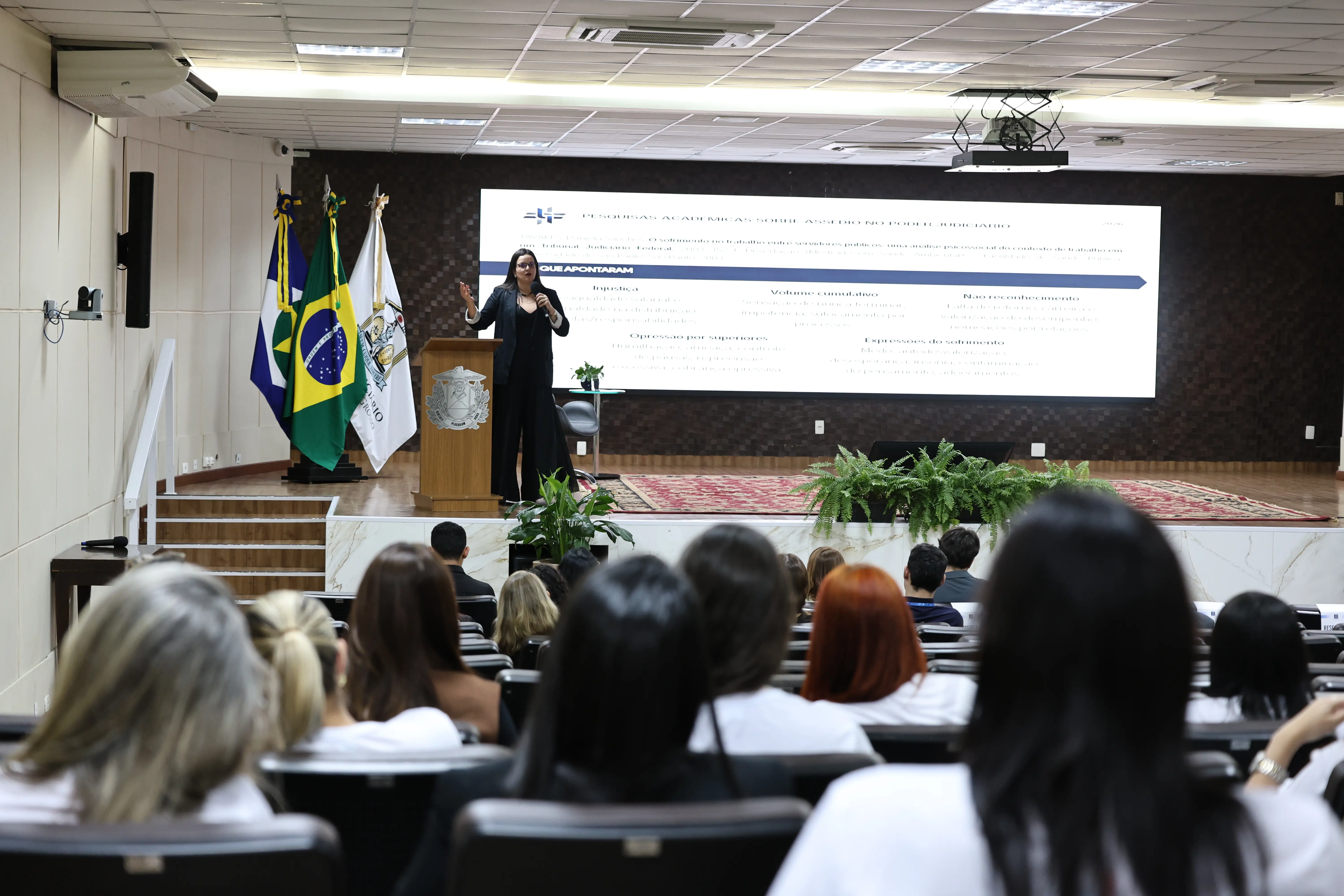 Uma mulher de cabelos escuros, vestindo roupa preta, gesticula enquanto fala ao microfone sobre um púlpito de madeira. Ao fundo, uma tela exibe slide sobre assédio no Poder Judiciário. Plateia numerosa assiste à palestra.