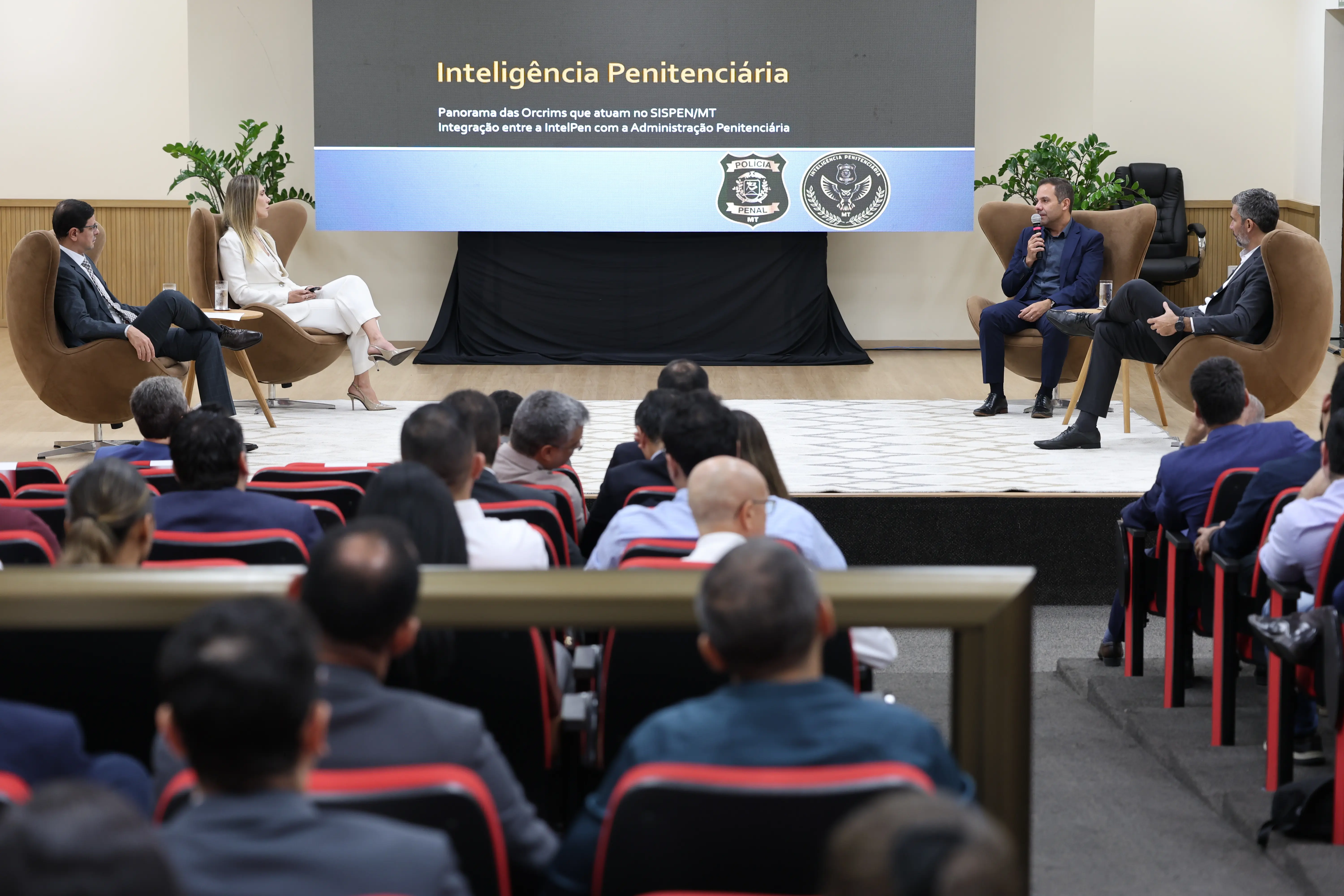 Quatro palestrantes debatem sentados em poltronas marrons no palco de um auditório. Ao fundo, um grande telão exibe o texto 