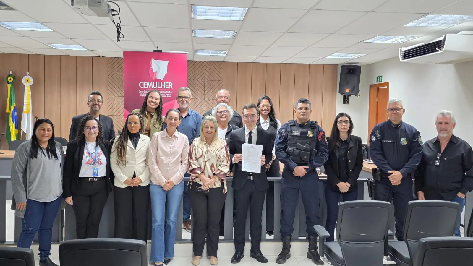 Grupo de cerca de 15 pessoas posa para foto em sala institucional. Ao centro, um homem de terno segura um documento. Ao fundo, banner rosa da CEMULHER e bandeiras do Brasil e de Mato Grosso.