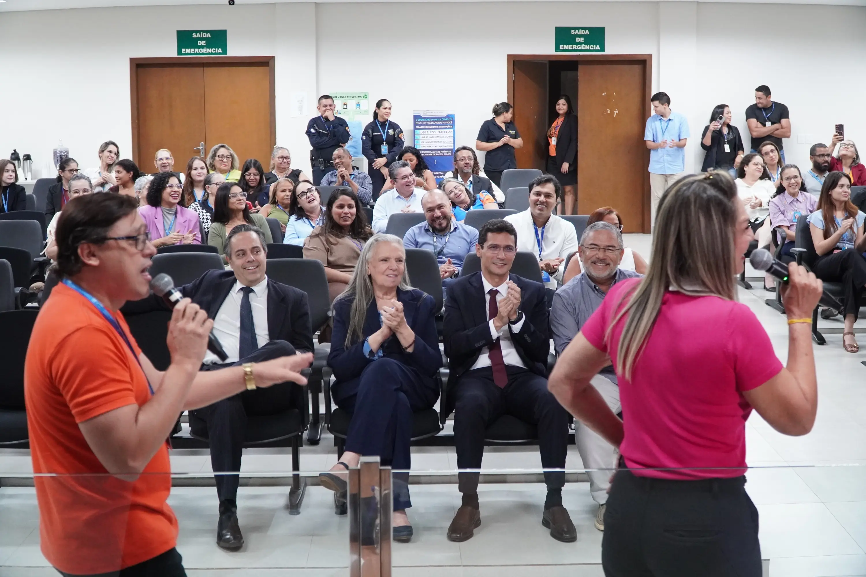 Foto que mostra um auditório lotado de pessoas sorridentes e aplaudindo. À frente, dois apresentadores com microfones — um de camiseta laranja e outro de blusa pink — animam o evento.