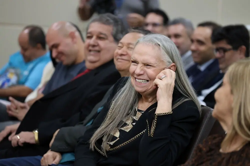 Foto horizontal que mostra a desembargadora Maria Erotides sentada na primeira fila da plateia do auditório, sorrindo. Ela é uma senhora branca, de longos cabelos lisos e grisalhos, usando blazer preto com detalhes dourados na gola. Ela está com a mão esquerda apoiando o rosto.