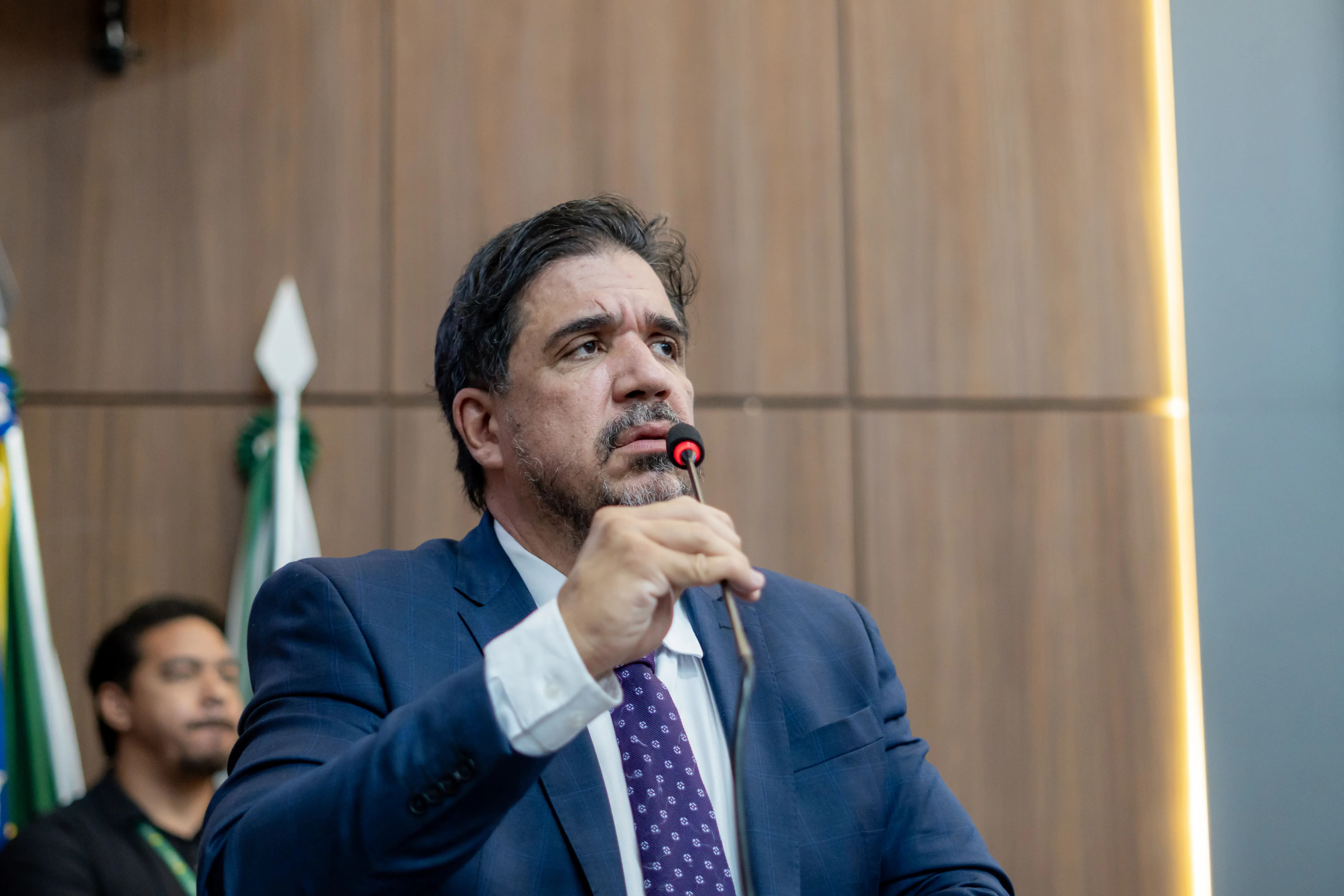 Homem de terno azul, camisa branca e gravata roxa fala ao microfone que segura com a mão direita. Ele tem cabelos escuros e barba por fazer, posicionado contra um painel de madeira.