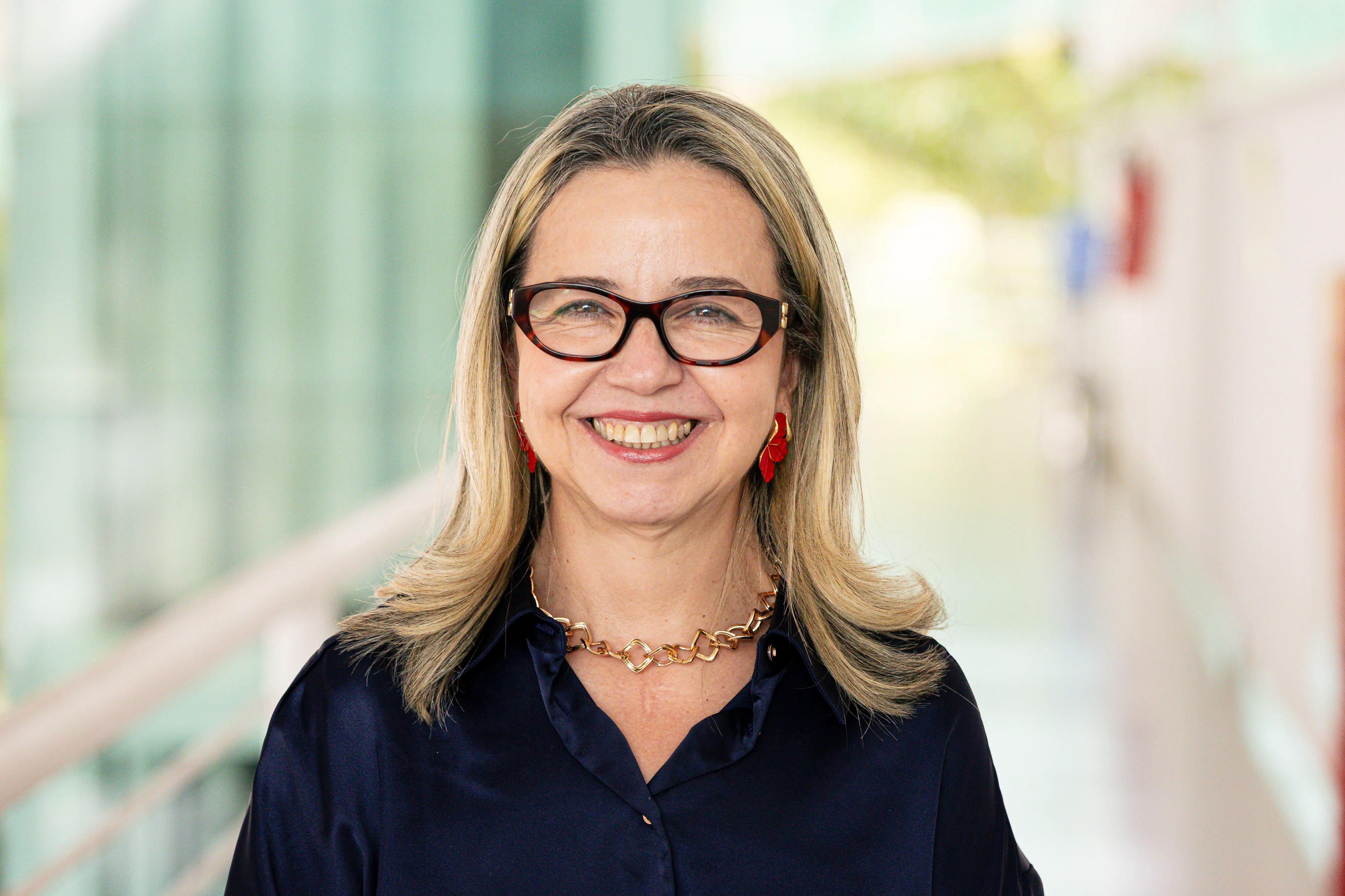 Mulher de pele clara sorrindo de frente, com óculos de armação escura, cabelos loiros lisos, colar de elos dourados e brincos vermelhos. Veste camisa azul-escura. Fundo de corredor desfocado.