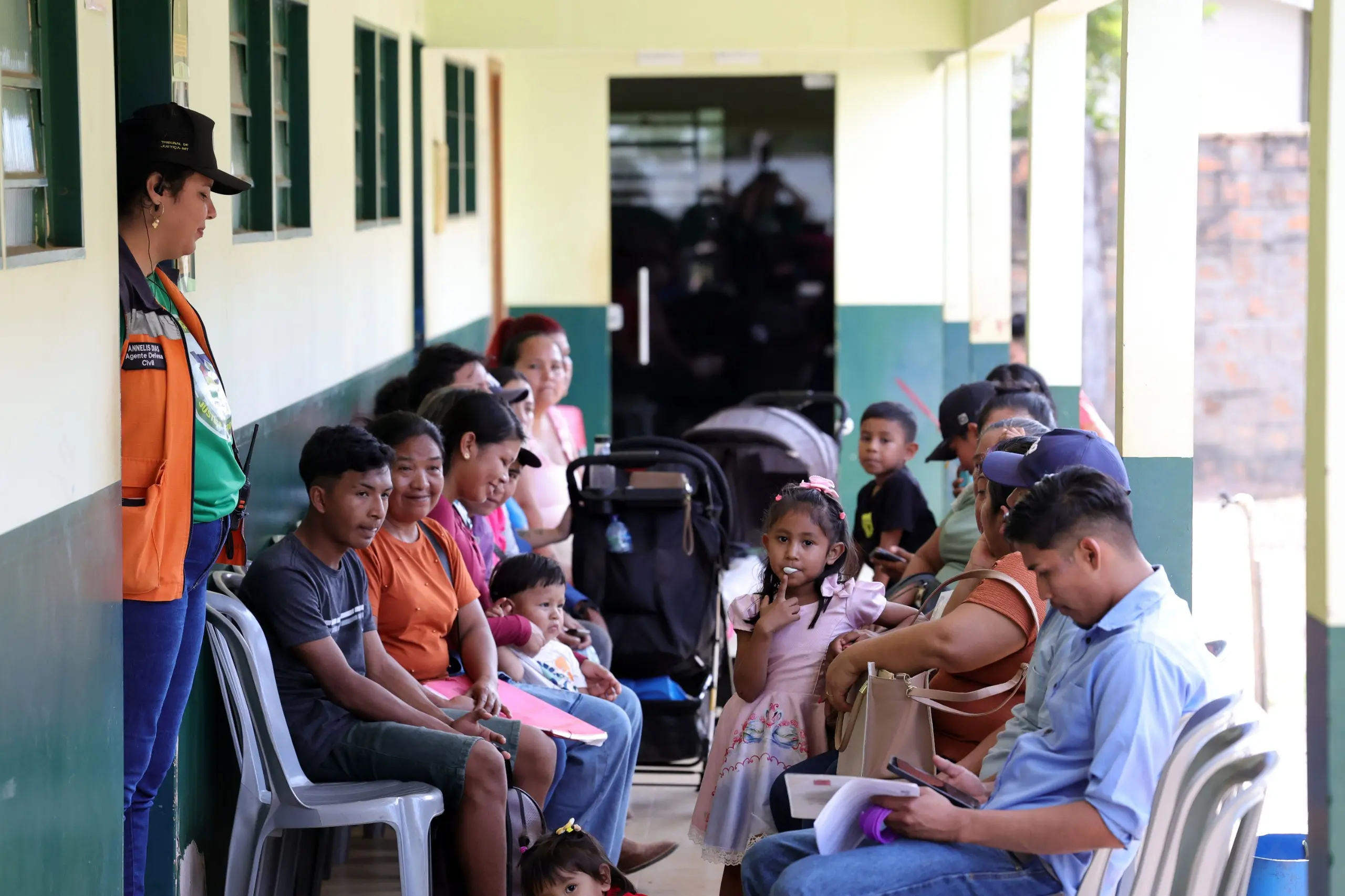 Foto horizontal em plano aberto que mostra o corredor de uma escola com paredes verdes de um lado e aberto para o pátio do outro, com várias pessoas sentadas, aguardando atendimento no mutirão Justiça Sem Fronteiras.