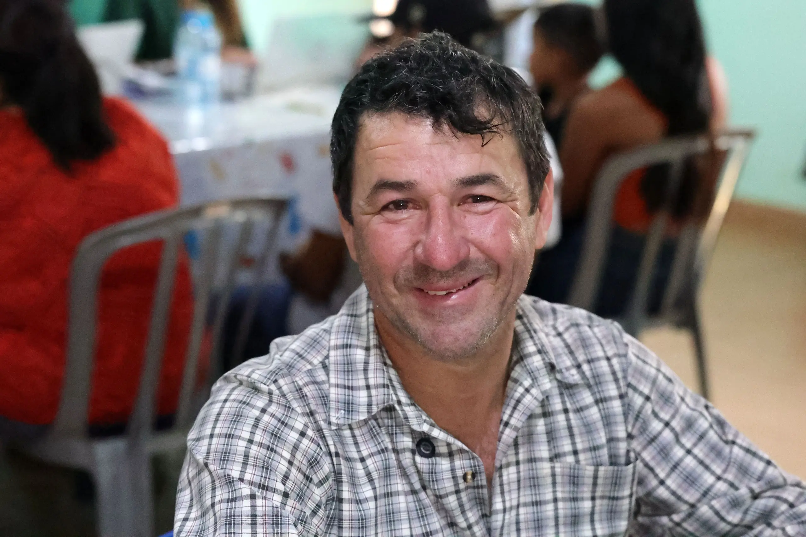 Homem de cabelos escuros e camisa xadrez sorri para a câmera. Ele está sentado em um ambiente interno e movimentado, com outras pessoas e mesas desfocadas ao fundo.