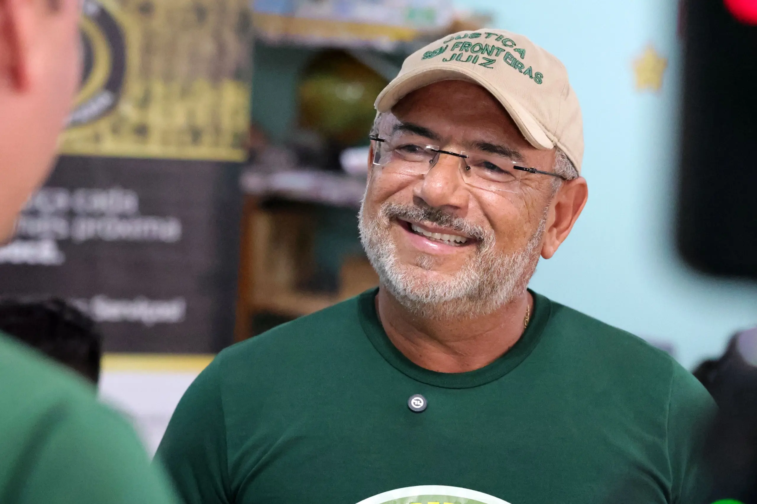 Homem sorridente de óculos, barba grisalha e camiseta verde. Ele usa um boné bege com a inscrição 