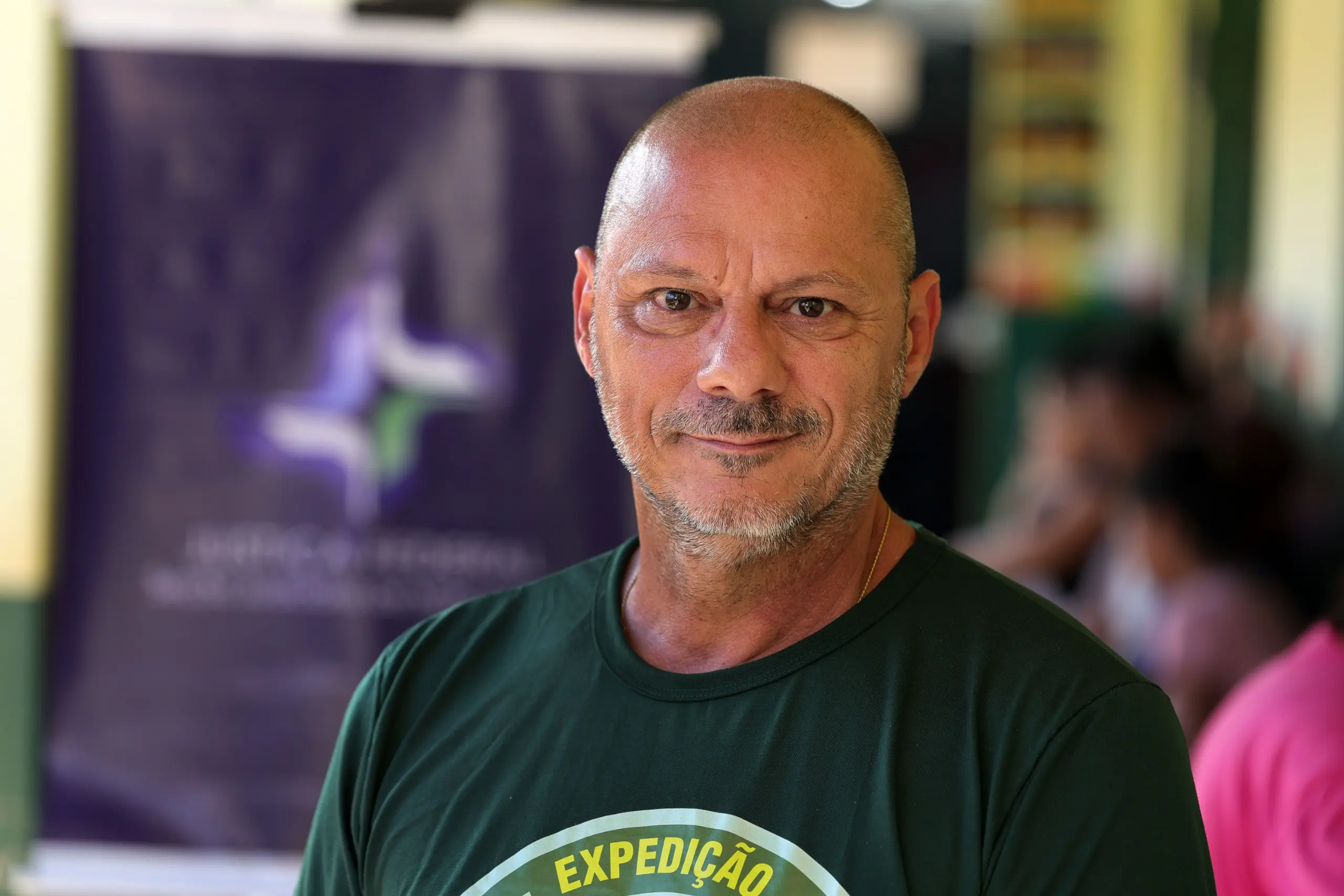 Homem com camiseta da Expedição Justiça Sem Fronteiras posa para fotografia em corredor ou área de atendimento, com pessoas desfocadas ao fundo.