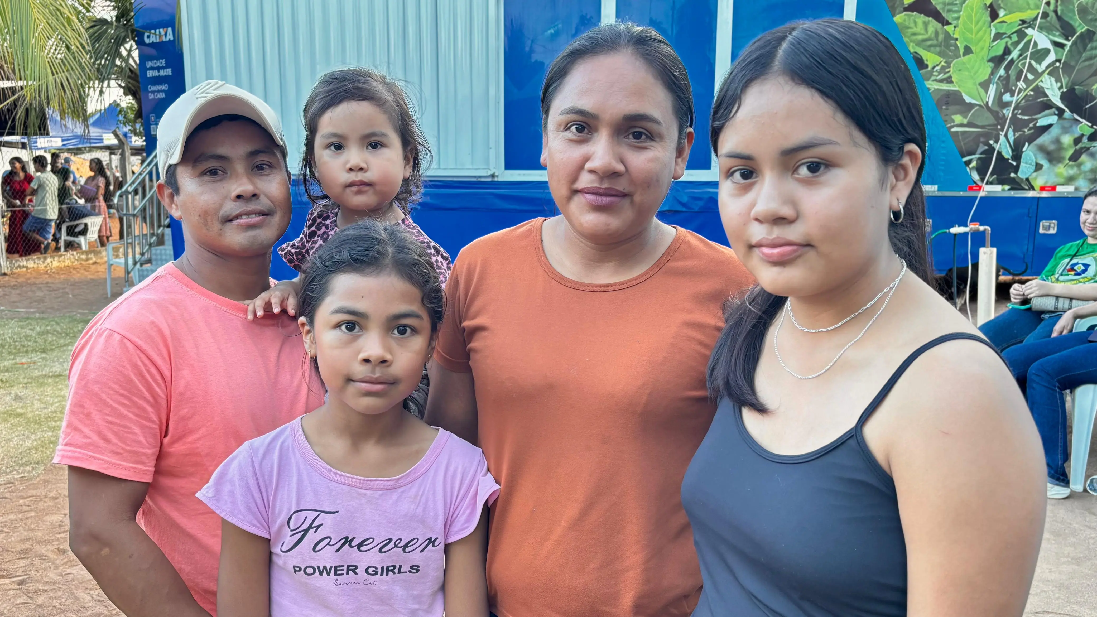 Uma família indígena de cinco pessoas posa junta ao ar livre diante de uma grande unidade móvel azul da Caixa Econômica Federal.