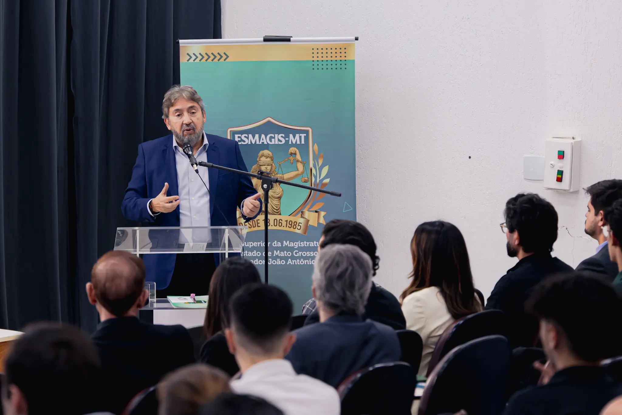 Um homem de terno azul discursa em um púlpito com microfone para uma plateia sentada. Ao fundo, há um banner verde com a inscrição ESMAGIS-MT.