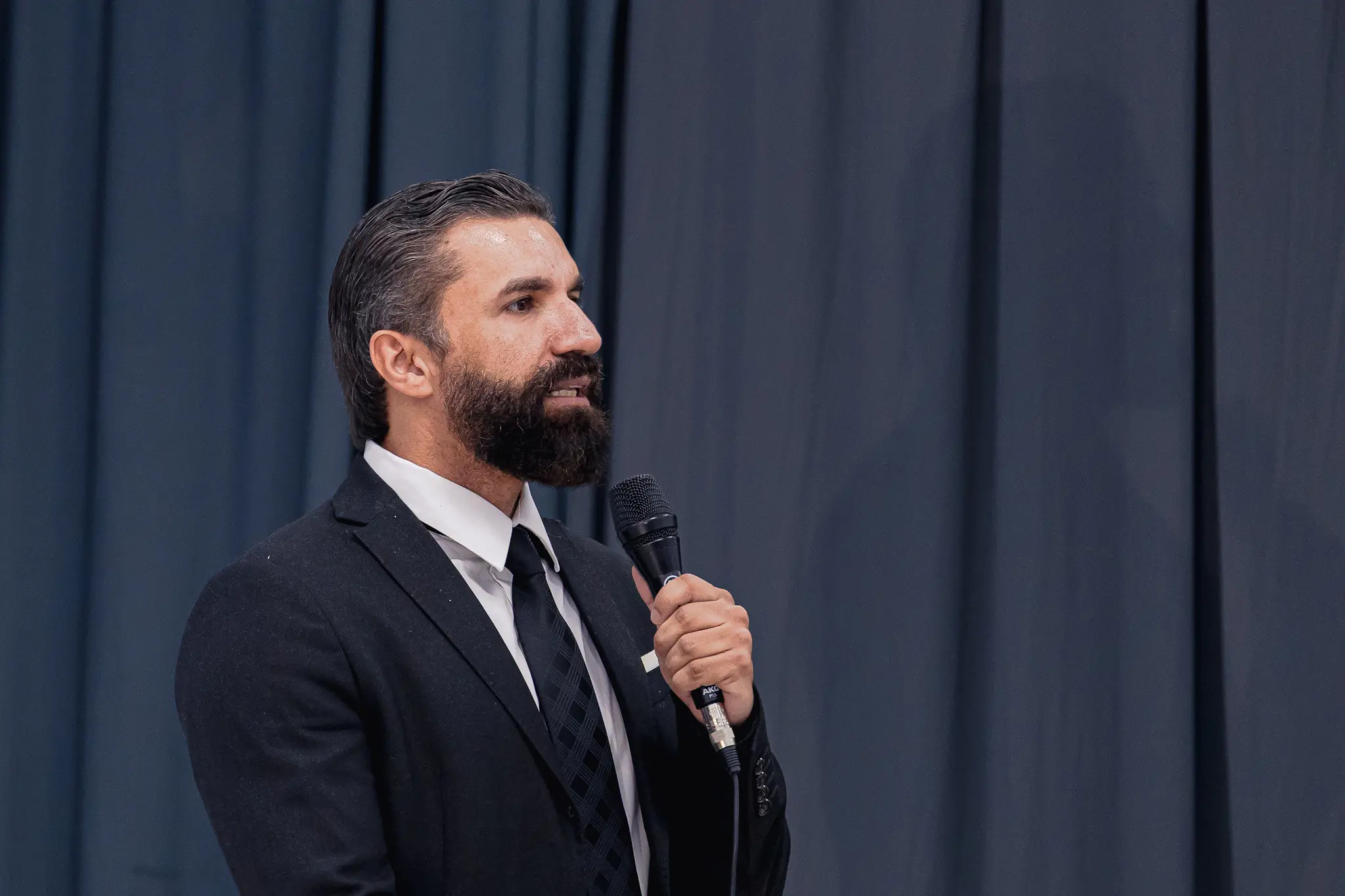 Homem de perfil, com barba e cabelo escuros, veste terno preto, camisa branca e gravata escura. Ele segura um microfone preto próximo à boca e fala, tendo uma cortina azul-escura ao fundo.