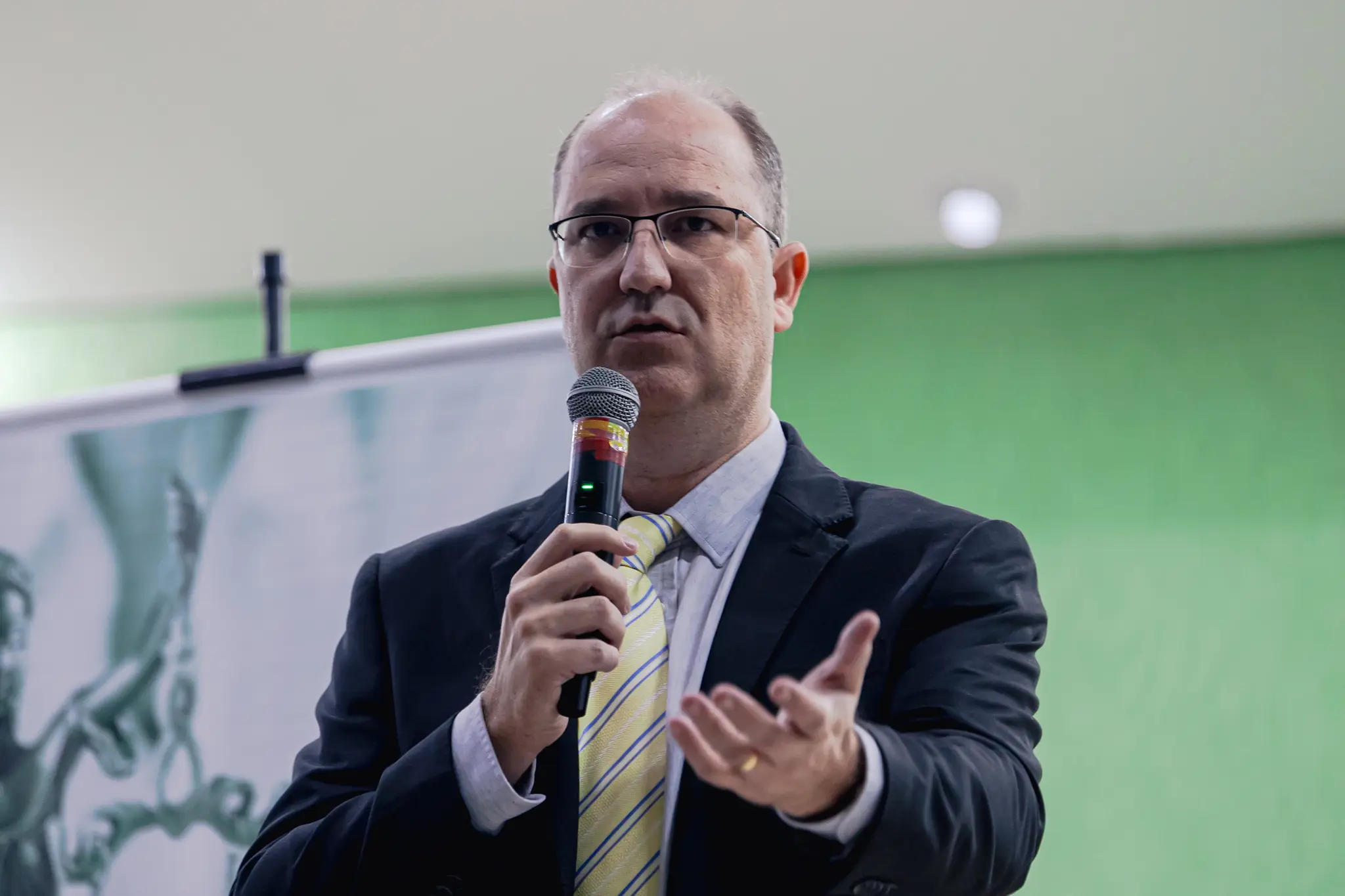 Homem de óculos fala ao microfone, gesticulando com a mão esquerda. Ele veste terno escuro, camisa clara e gravata amarela listrada. Ao fundo, uma parede verde e um banner com a deusa da justiça.