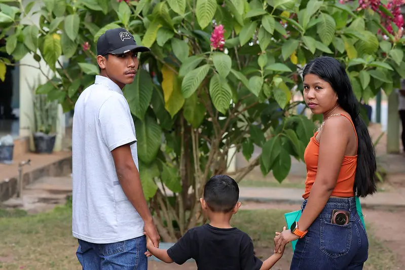 Casal indígena de costas, segurando as mãos de uma criança pequena, olha para trás. Ao fundo, um grande arbusto verde com flores rosas. Ambiente ao ar livre.