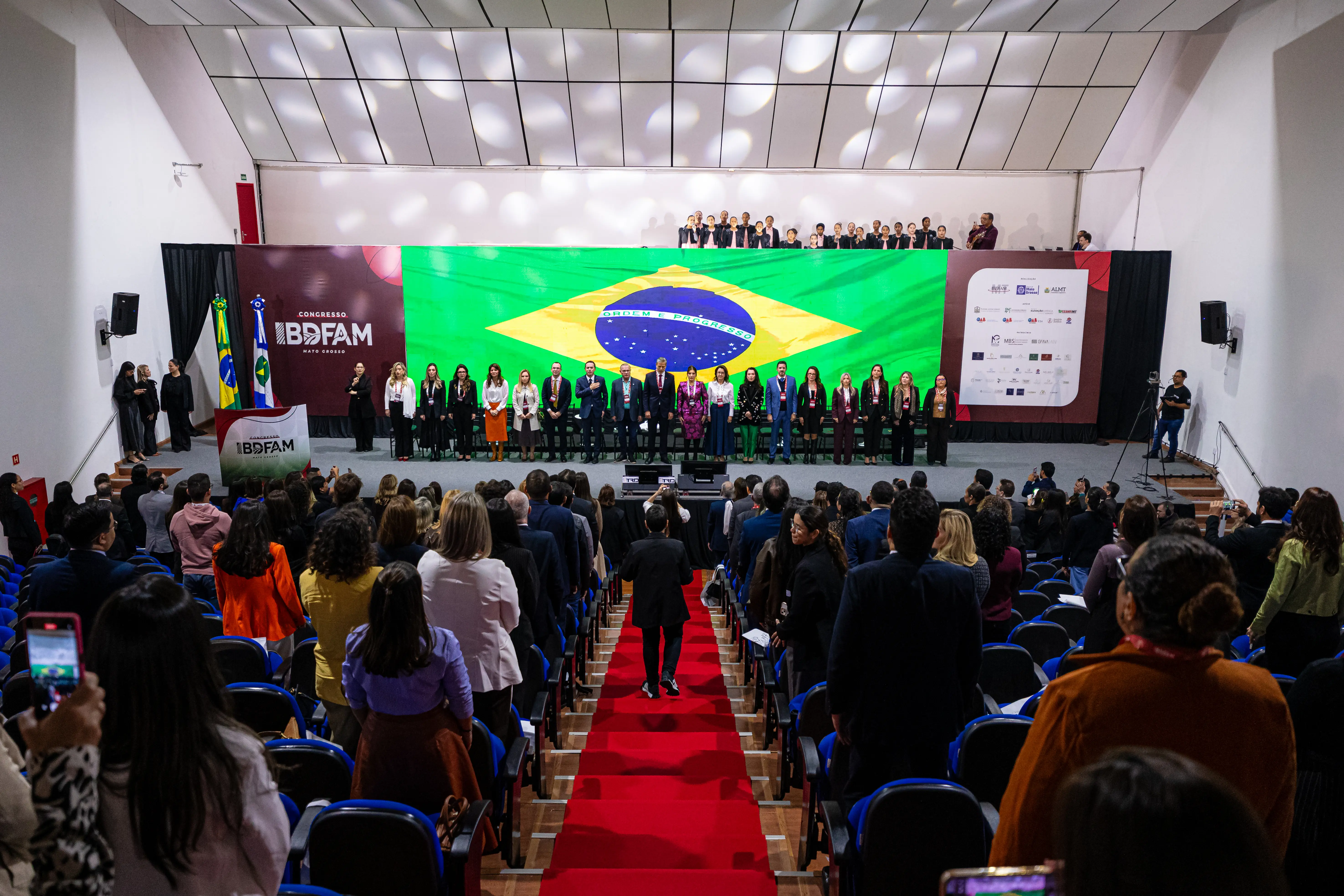 Visão geral de um auditório lotado com pessoas de pé. No palco iluminado, autoridades perfiladas diante de um grande painel com a bandeira do Brasil. Um tapete vermelho cruza o corredor central.