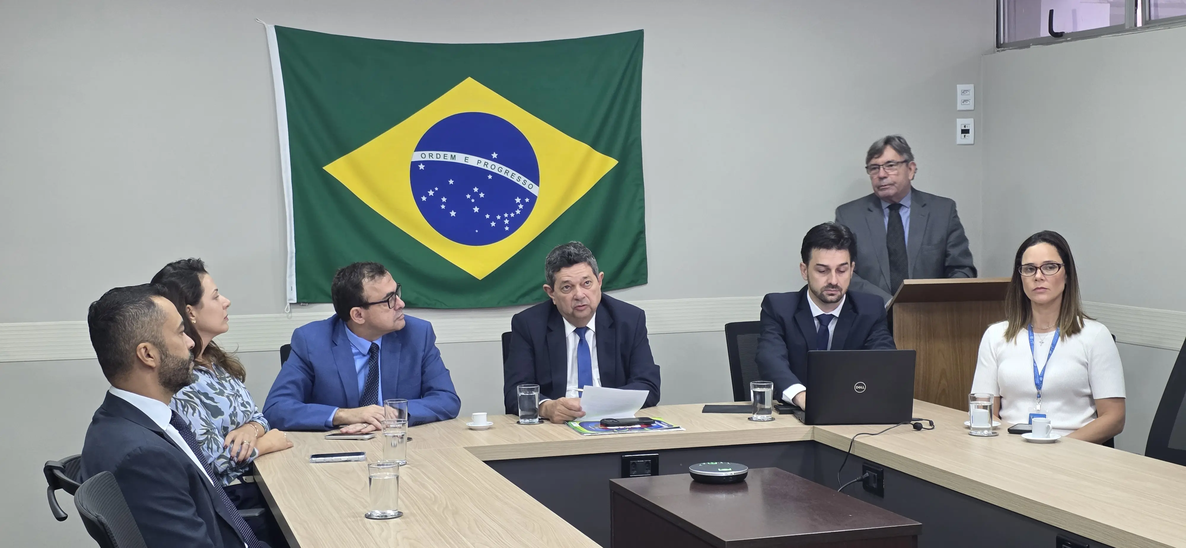 Reunião institucional com seis pessoas em torno de uma mesa de madeira em formato de U. Ao fundo, a bandeira do Brasil na parede. O homem ao centro fala enquanto segura um documento.