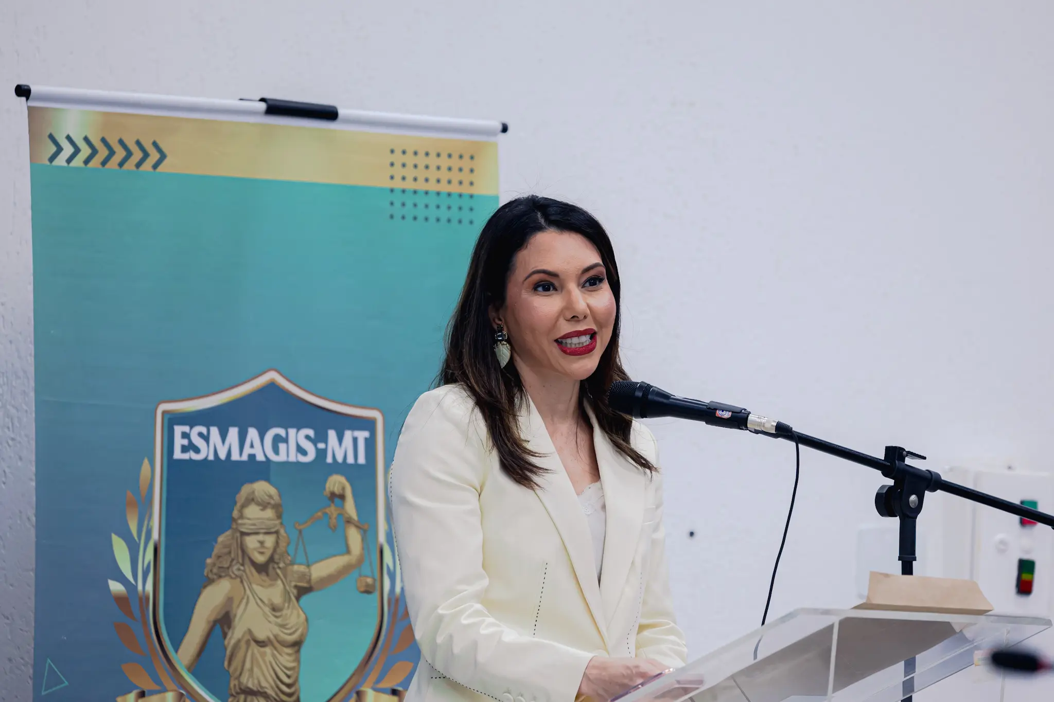 Mulher de cabelos escuros e blazer claro sorri ao falar num microfone em púlpito acrílico. Ao fundo, parede branca e um banner verde com a imagem da deusa da justiça e o logotipo ESMAGIS-MT.
