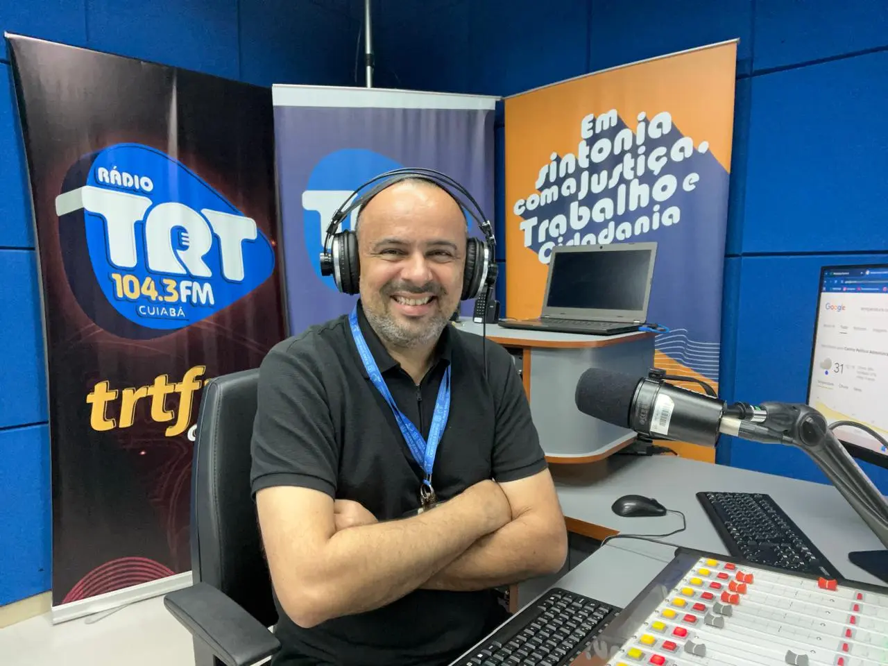 Homem sorridente de pele clara e com barba rala sentado em estúdio de rádio com braços cruzados e fone de ouvido. À sua frente, há uma mesa de som, teclado e microfone. Ao fundo, painéis azuis e banners com logotipos da