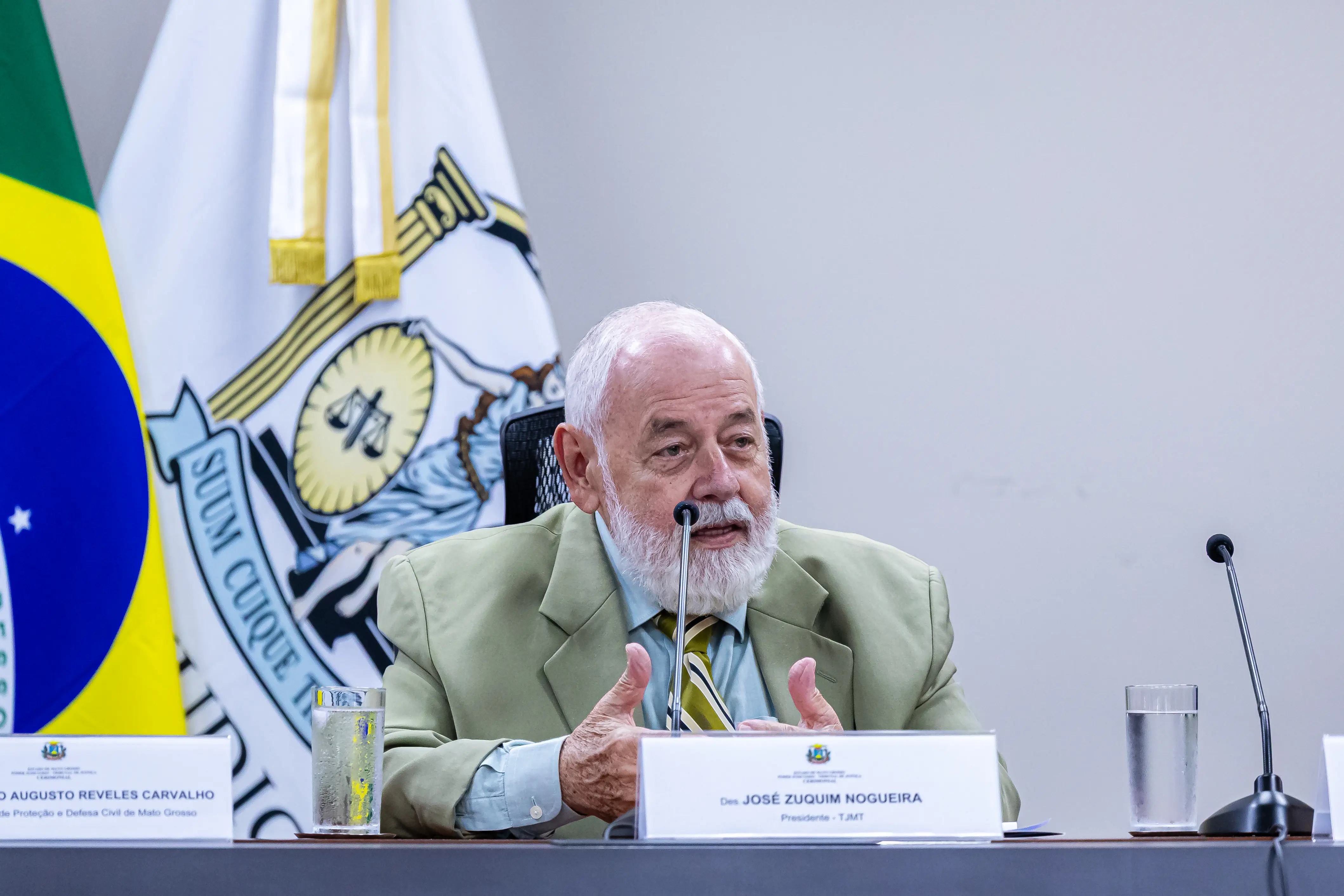 Homem de barba branca, vestindo paletó verde-claro e gravata listrada, fala ao microfone instalado em uma bancada com placas de identificação. Ao fundo, as bandeiras do Brasil e do Judiciário