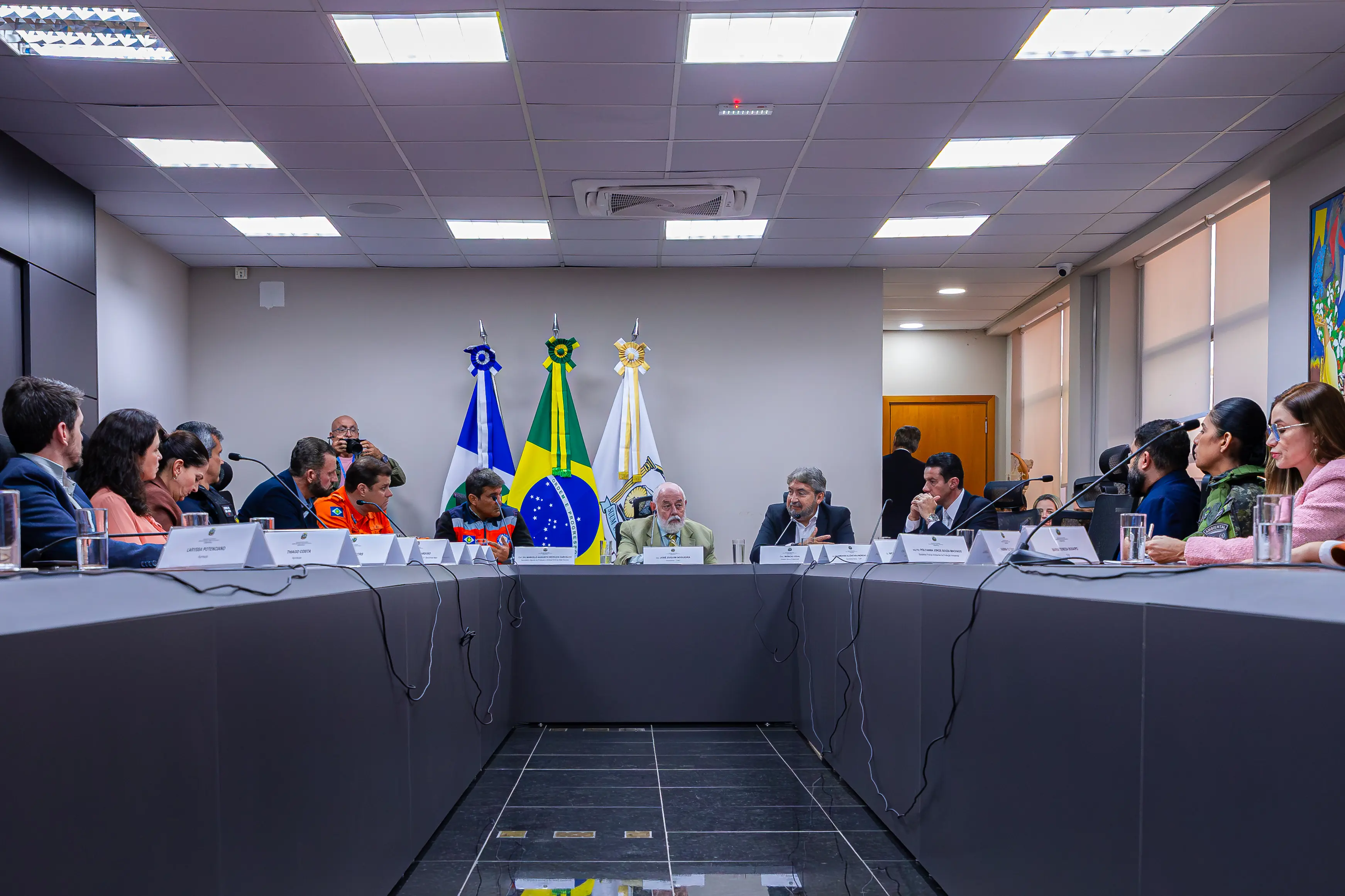 Autoridades reunidas ao redor de uma grande mesa de conferência em U. Ao fundo, posicionadas ao centro, estão as bandeiras de Mato Grosso, do Brasil e do Judiciário.