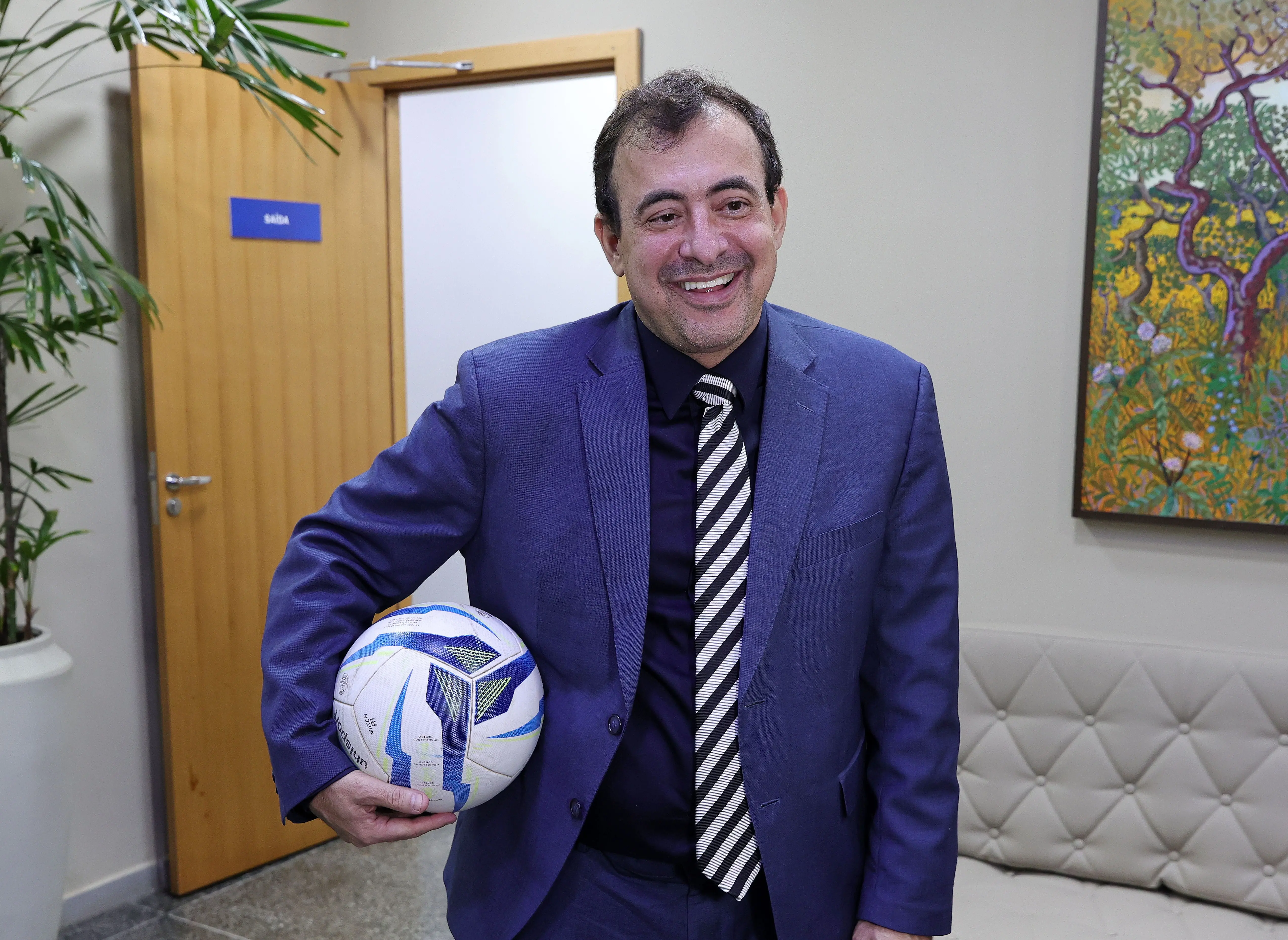 Foto horizontal que mostra o juiz Jorge Alexandre Martins Ferreira da cintura pra cima, em pé, sorrindo e segurando uma bola de futebol. Ele é um homem branco, de cabelos castanhos, usando camisa azul marinho, terno azul e gravata listrada.