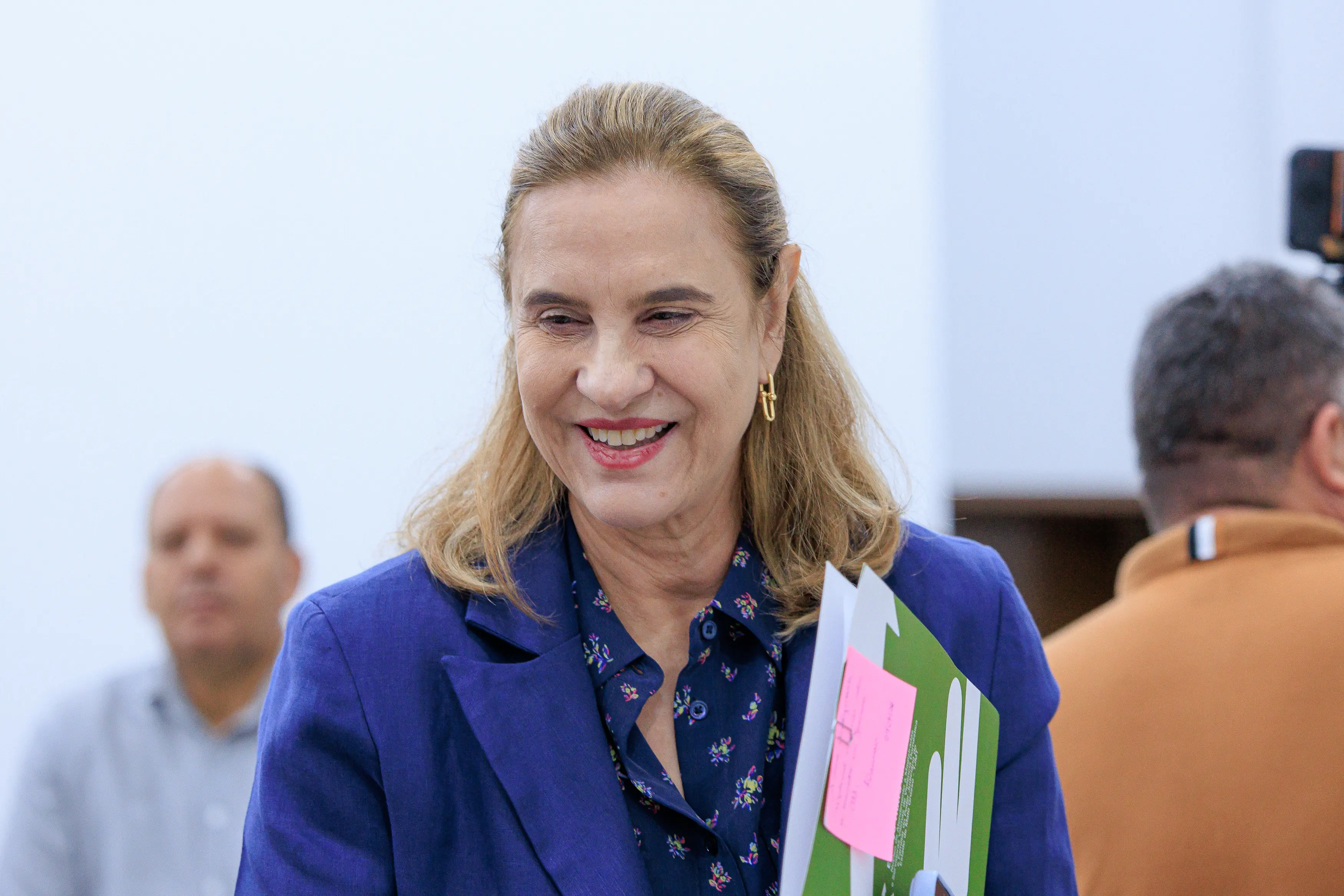 Desembargadora Vandymara Galvão sorri e caminha, segurando uma pasta. Ela veste um blazer azul-marinho e uma blusa azul estampada. O fundo é composto por um ambiente interno claro com duas outras pessoas desfocadas.