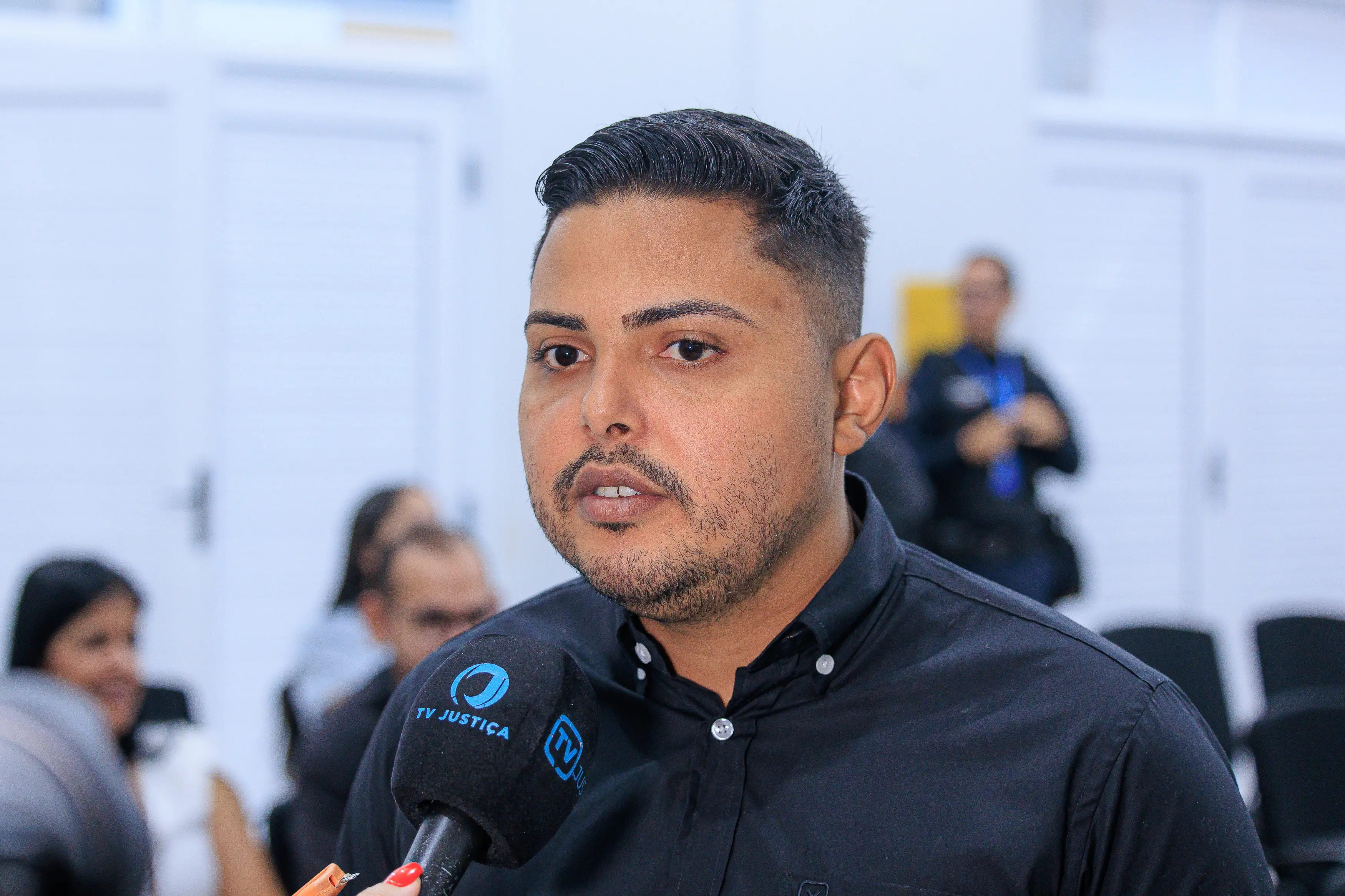 Secretário municipal de Assistência Social de Jaciara, Renan Henrique Ribeiro é jovem de pele morena e cabelo curto e escuro, cortado em estilo degrade, fala para um microfone com o logotipo