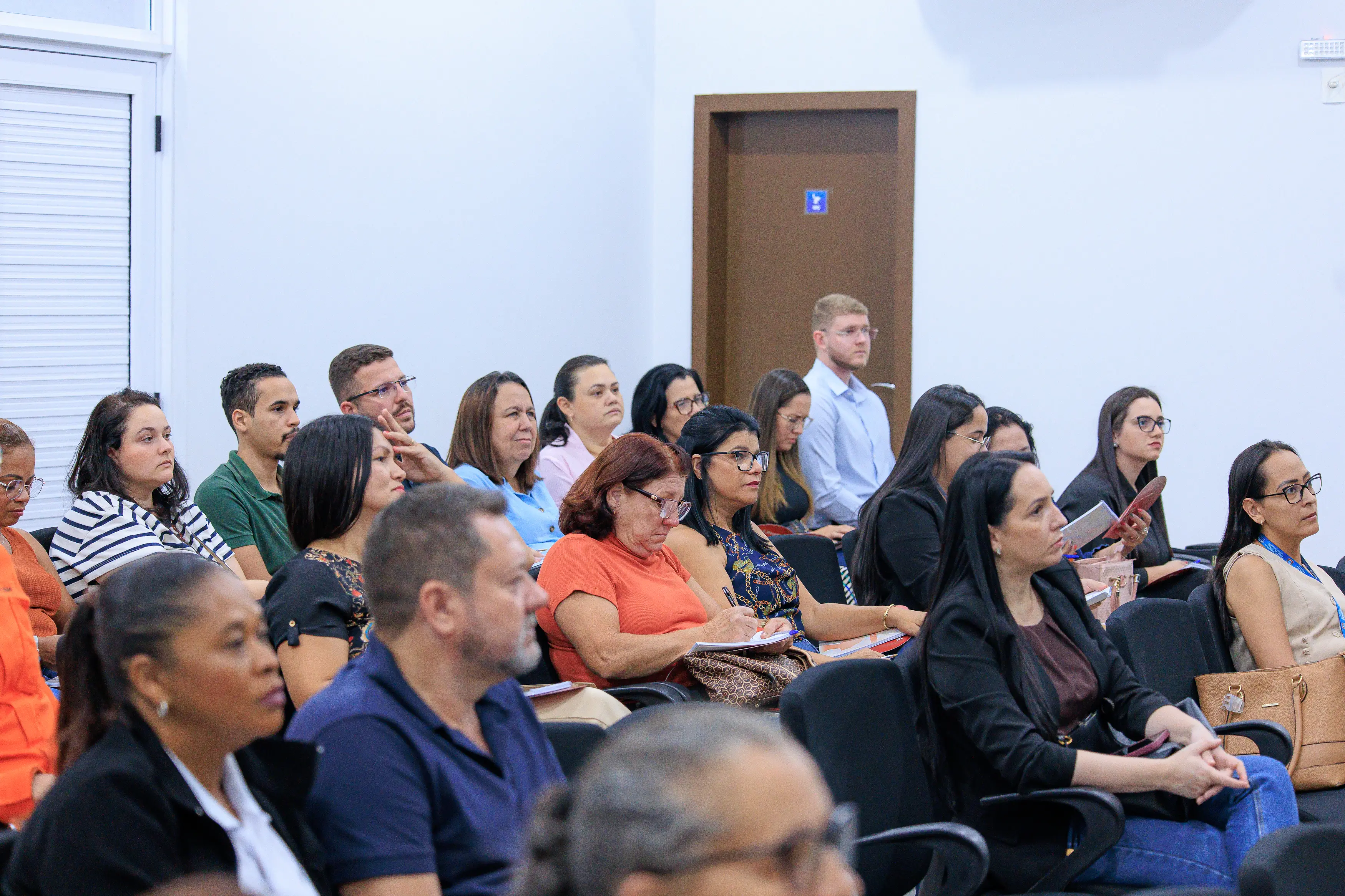 Um grupo de cerca de vinte pessoas está sentado em filas de cadeiras pretas em um auditório interno claro. A maioria das pessoas está olhando para a frente, em direção a um local fora do quadro. Várias delas seguram cadernos ou bolsas. No fundo, há uma porta de madeira.