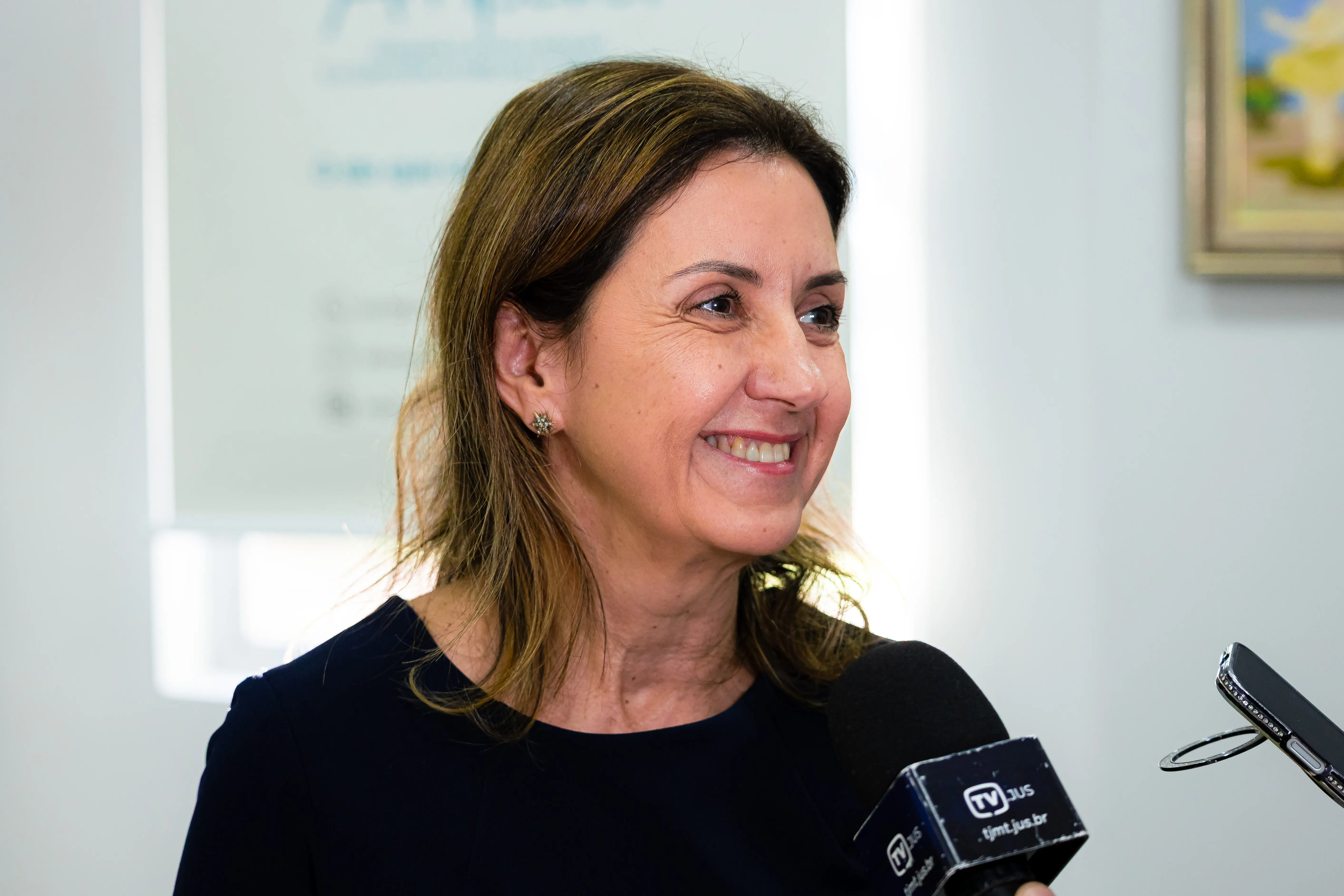 Mulher de cabelos castanho-claros sorri enquanto fala ao microfone da TV JUS. Veste blusa escura e brinco pequeno. Ao fundo, banner institucional desfocado.