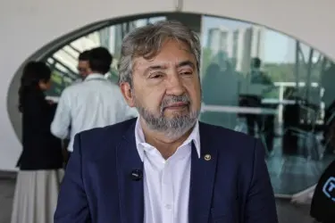 Homem com barba grisalha veste paletó azul e camisa branca, em ambiente interno com vidro ao fundo; outras pessoas aparecem desfocadas ao redor, sugerindo evento ou entrevista.