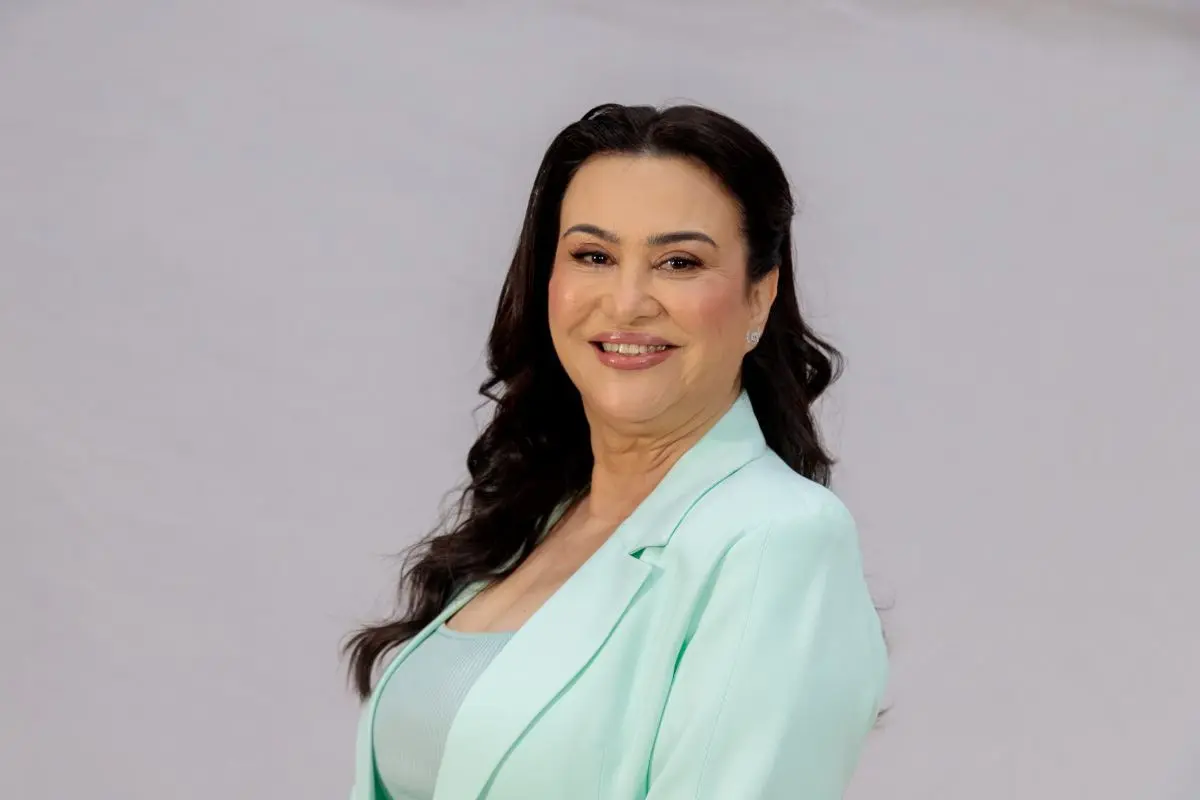 Mulher de longos cabelos escuros ondulados, sorrindo de frente para a câmera. Ela veste um blazer verde-água sobre uma blusa de tom semelhante, com fundo cinza claro e neutro.