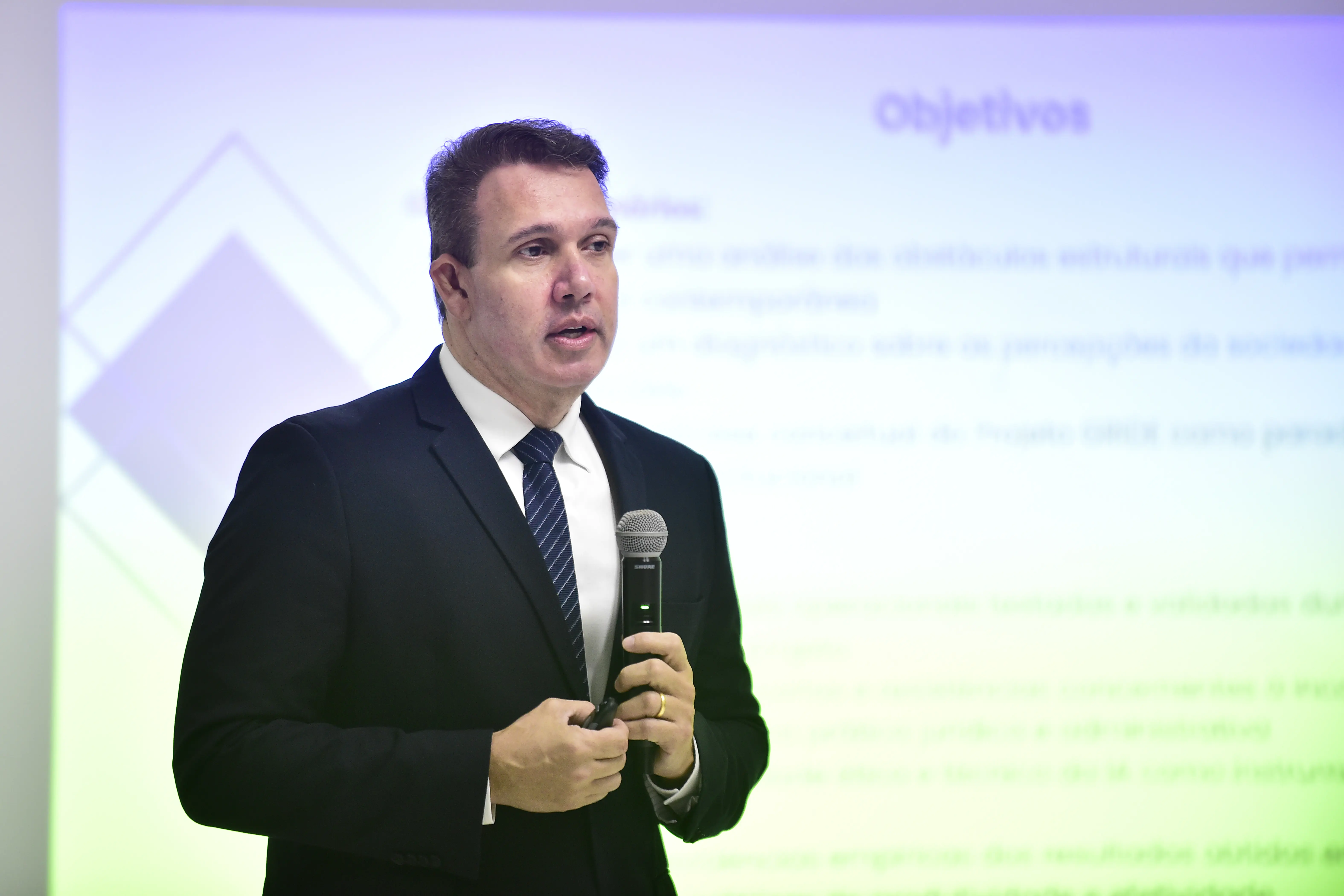 Desembargador Luiz Octávio Saboia Ribeiro em um terno escuro e gravata listrada, está em pé e fala em um microfone, com uma projeção ao fundo.